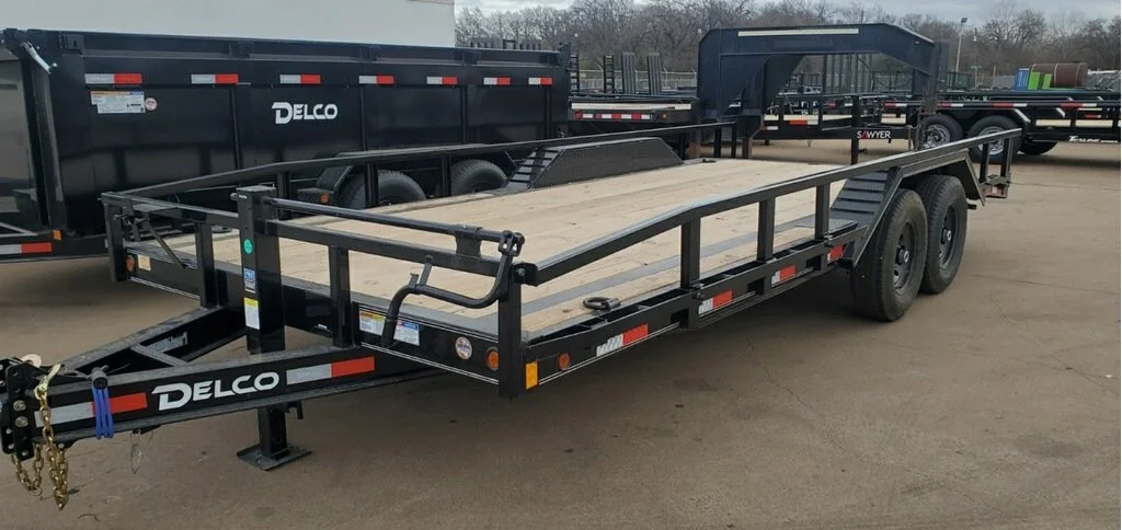 A flatbed utility trailer with a black metal frame and wooden deck in an outdoor lot. The trailer has two axles with large tires, safety railings on the sides, and a hitch for towing.