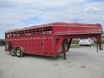 Red livestock or horse trailer parked outdoors on gravel, with a white vehicle visible in the background under a blue sky with clouds.