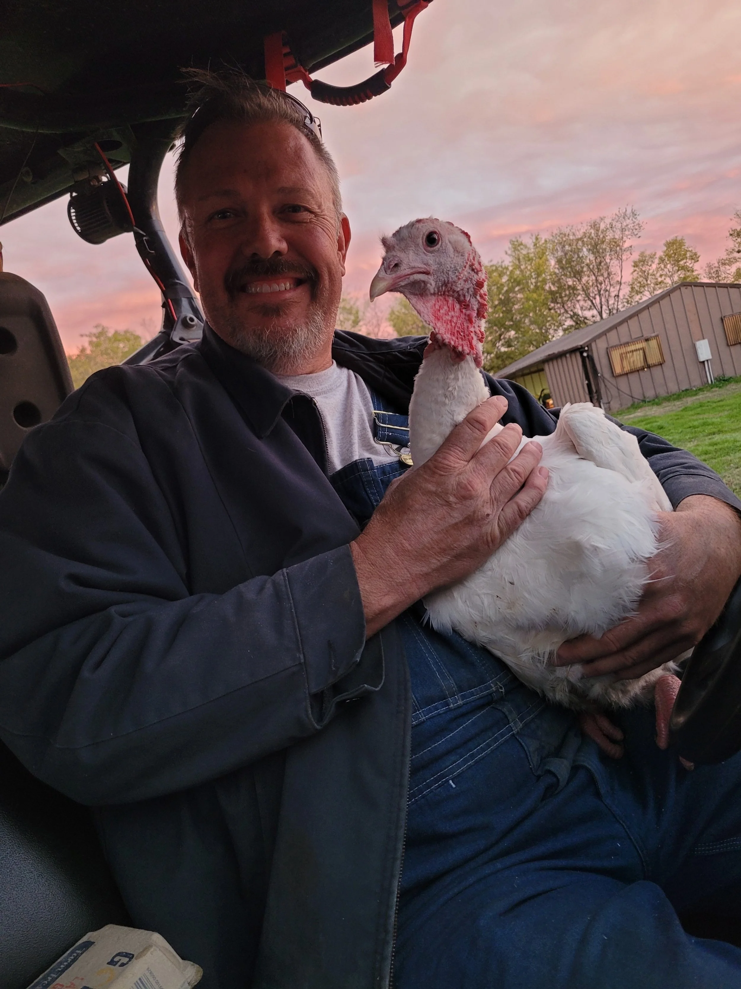 A man smiling while sitting on a tractor, holding a white turkey with a red head, during sunset outdoors.