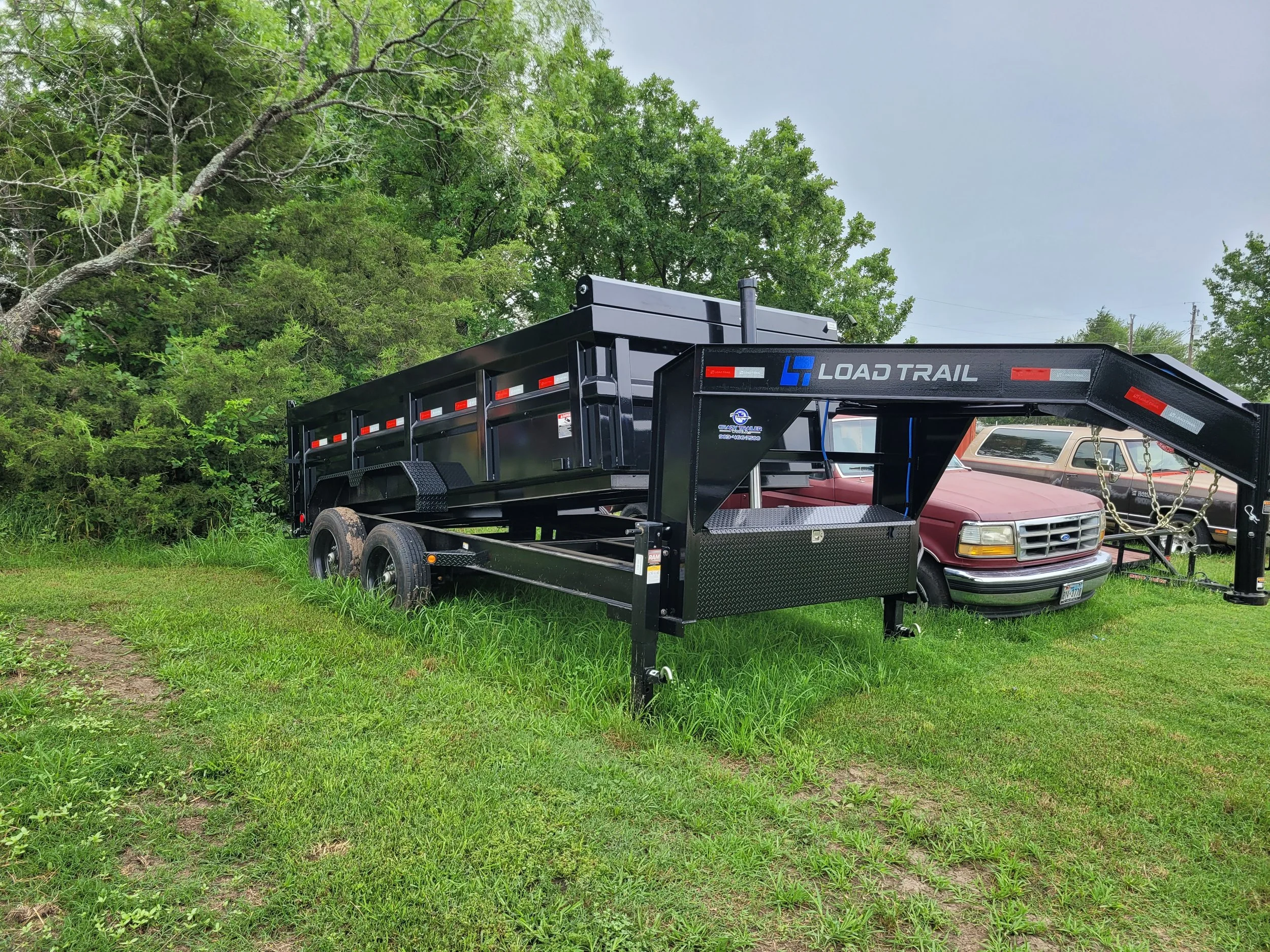 Black Load Trail flatbed trailer parked on grassy area beside a red pickup truck and other vehicles, with trees in the background.