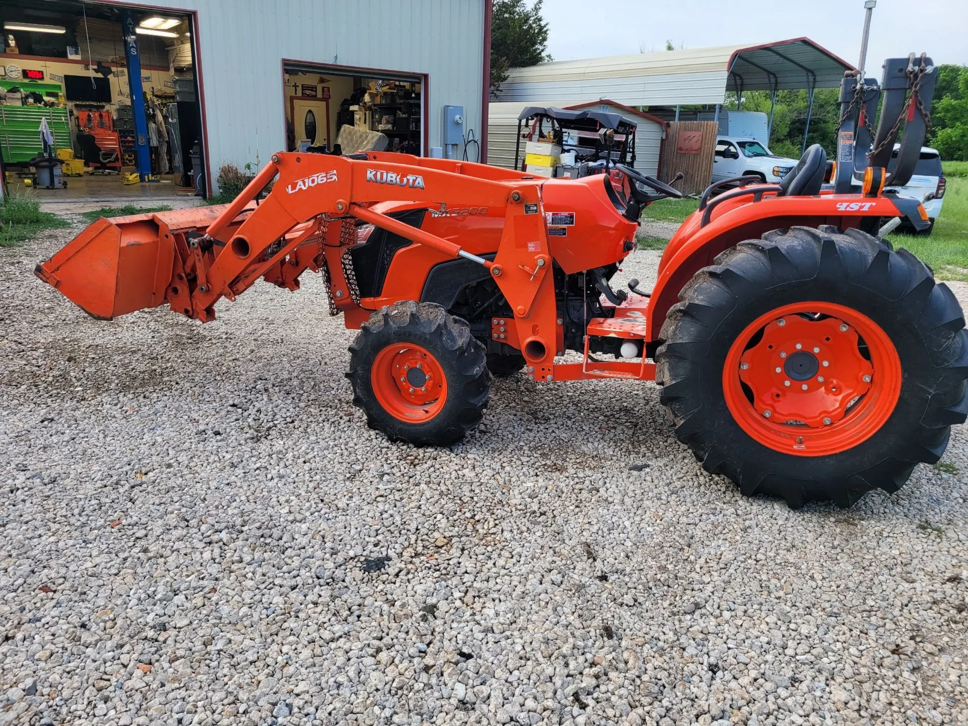 Orange Kubota tractor with front loader attachments parked on gravel outside a workshop with open garage doors. Other vehicles and structures are visible in the background.