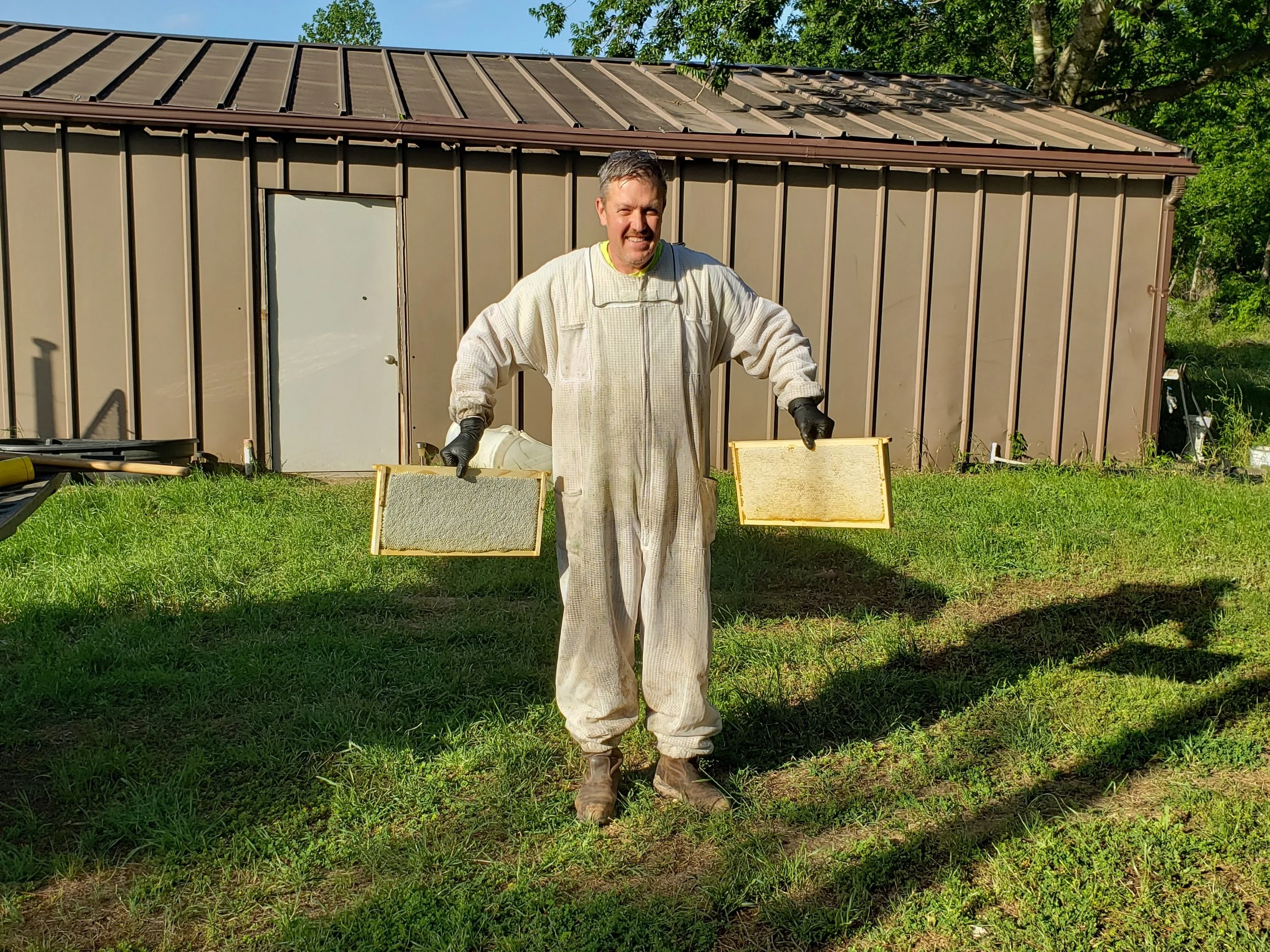 A man wearing protective clothing and gloves holding two beekeeping frames outdoors on a grassy area with a brown shed in the background.