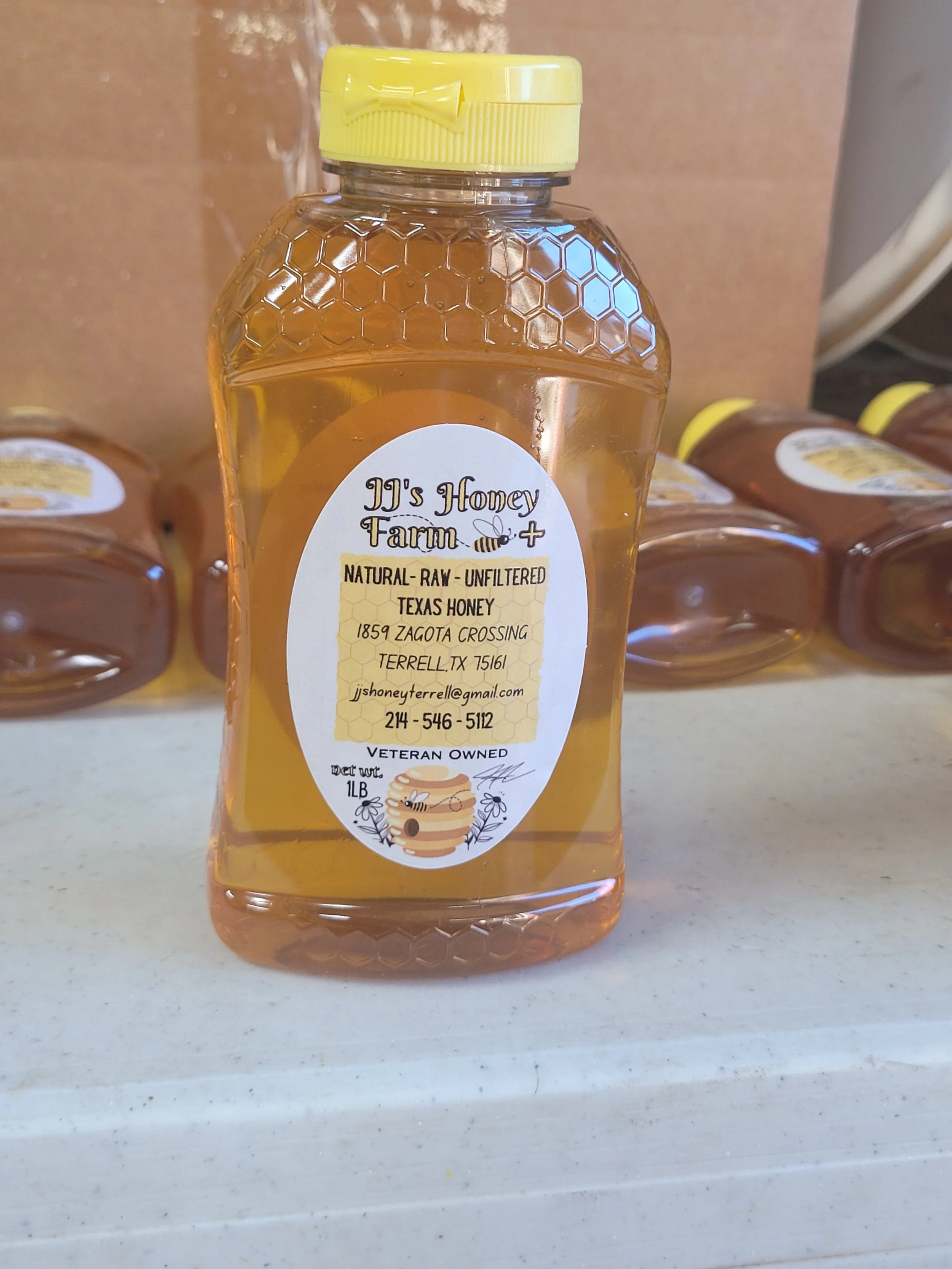 Bottle of raw, unfiltered Texas honey from JJ's Honey Farm with a yellow lid and a honeycomb pattern on the top part of the bottle, placed on a white surface with other honey bottles in the background.
