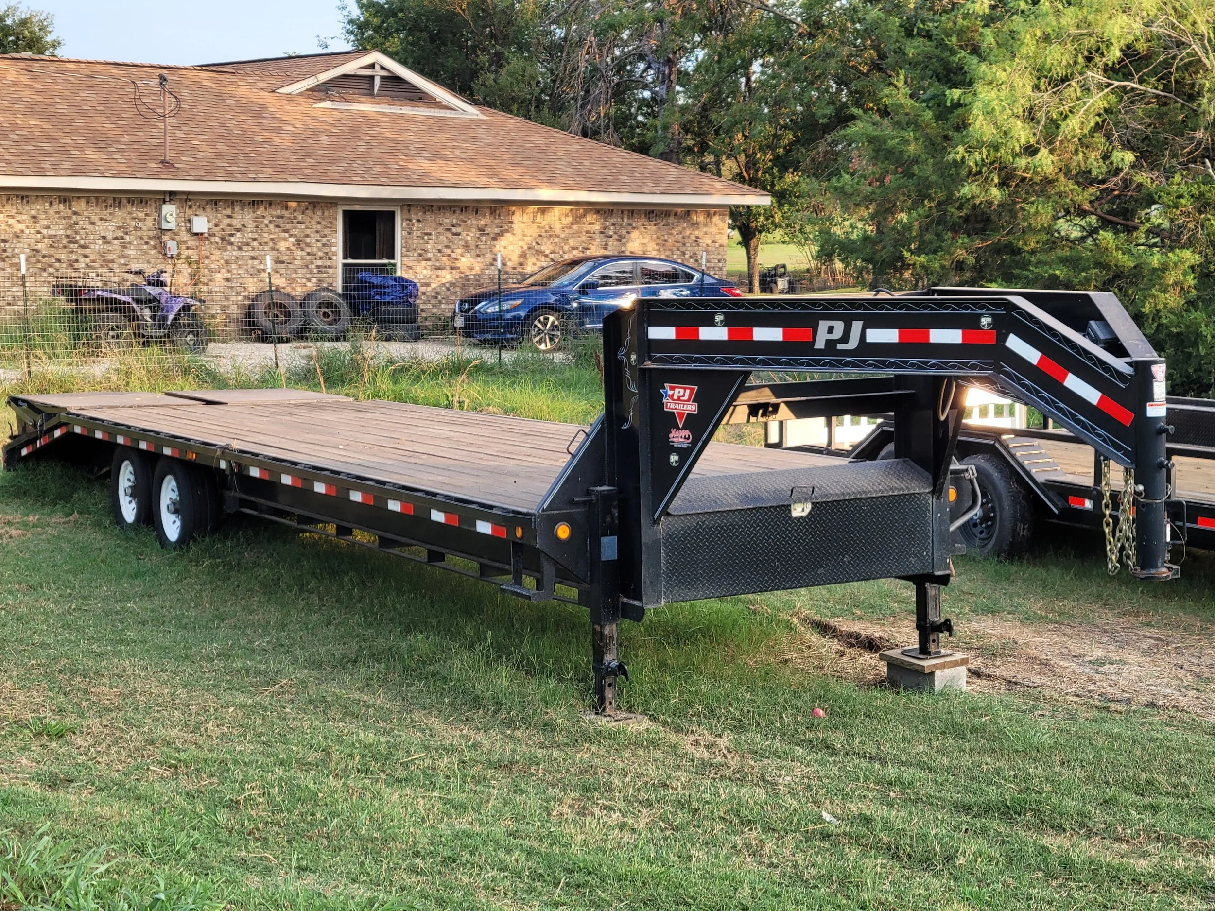 A black flatbed trailer with a wooden deck parked on grass in front of a house, with a car and a purple ATV in the background.