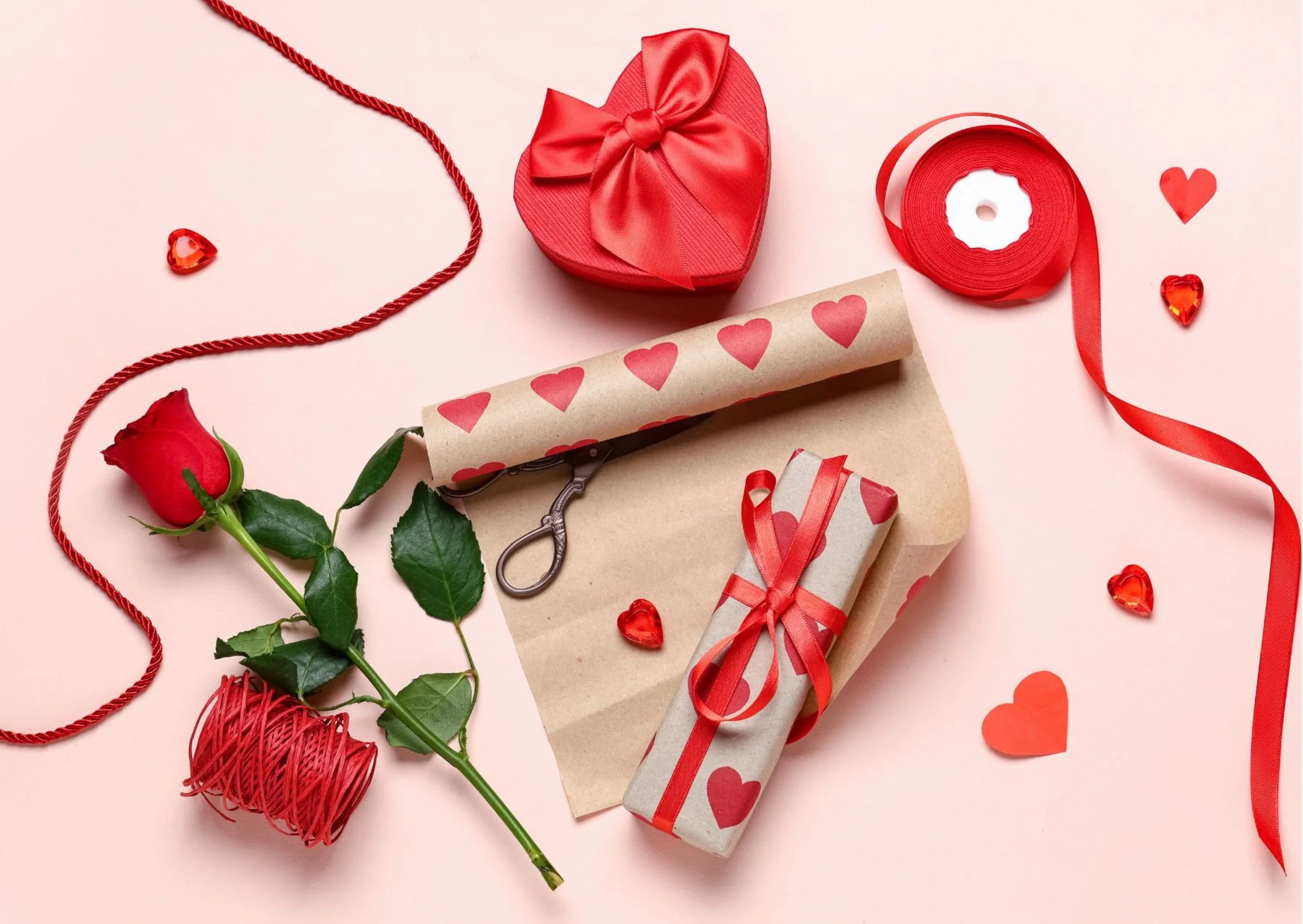 Valentine's Day gift wrapping supplies including a red heart-shaped box with a satin bow, a roll of red ribbon, a small wrapped gift with a red ribbon, a red rose, a spool of red thread, and heart-shaped decorations on a pink background.