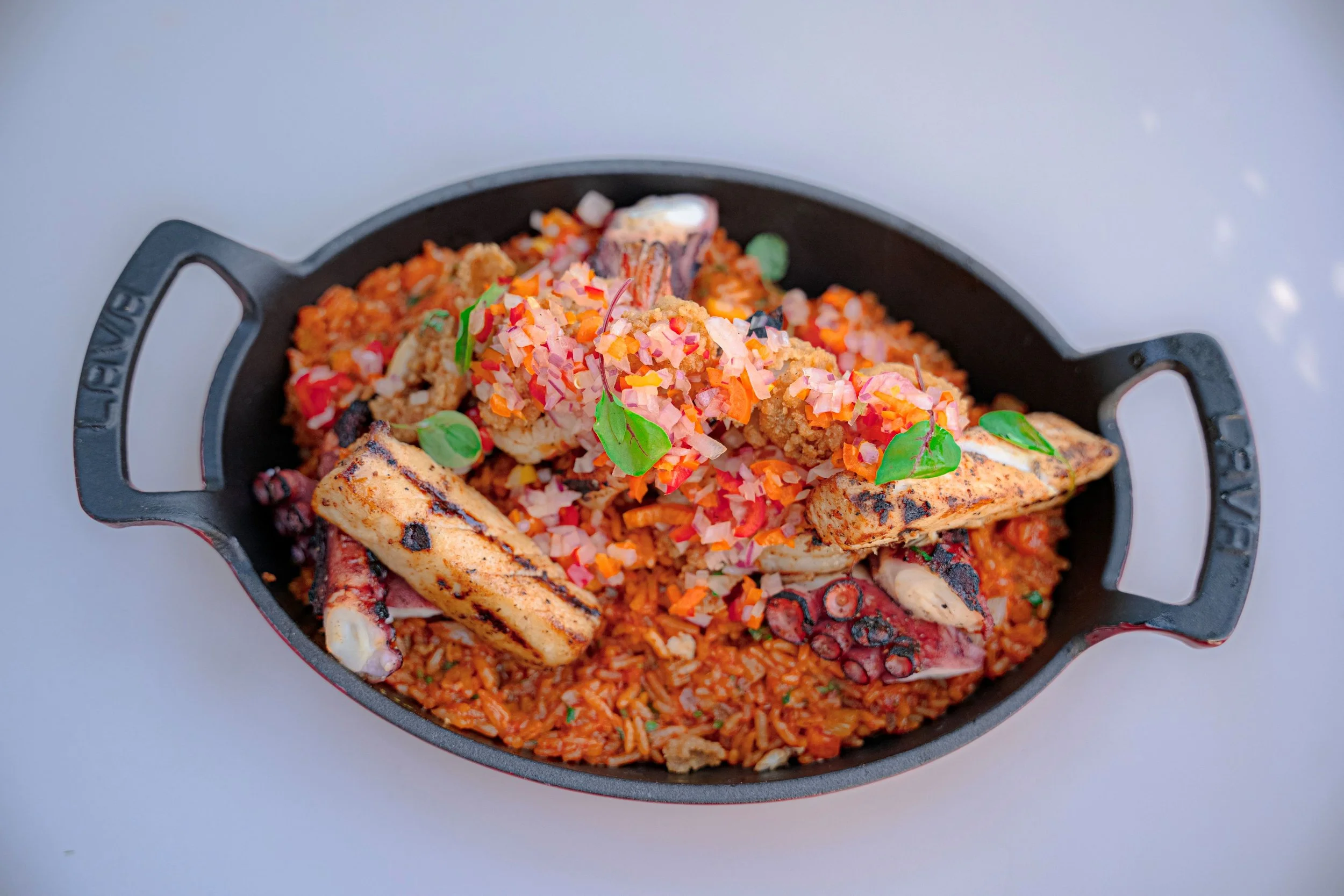 Seafood paella with grilled seafood, vegetables, and herbs in a black serving dish.