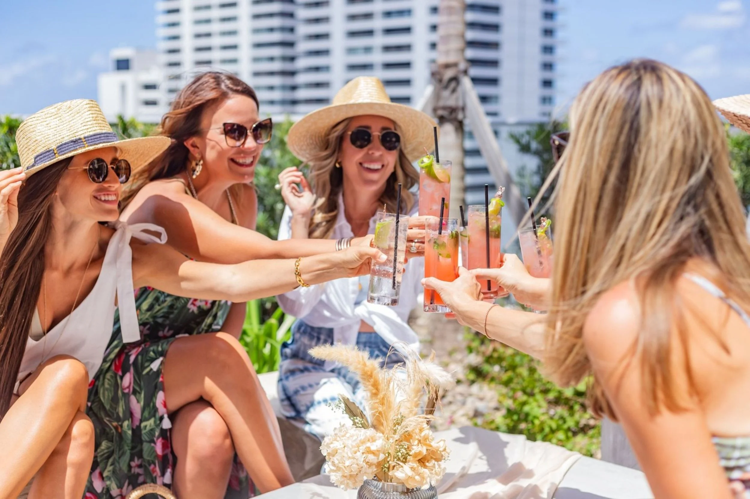 Four women on a rooftop deck celebrating with drinks, wearing summer clothing and sun hats, smiling and toasting each other, with city buildings in the background.