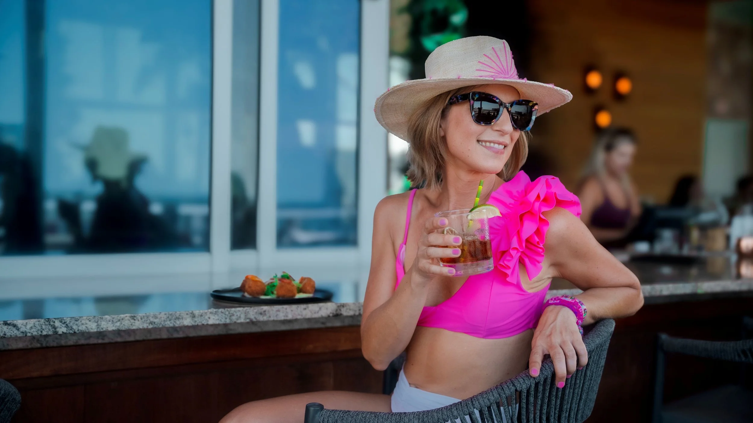 Woman wearing a large sunhat and sunglasses, holding a drink with a lime in a bright pink top with ruffle details, sitting at a bar with a plate of food in the background.