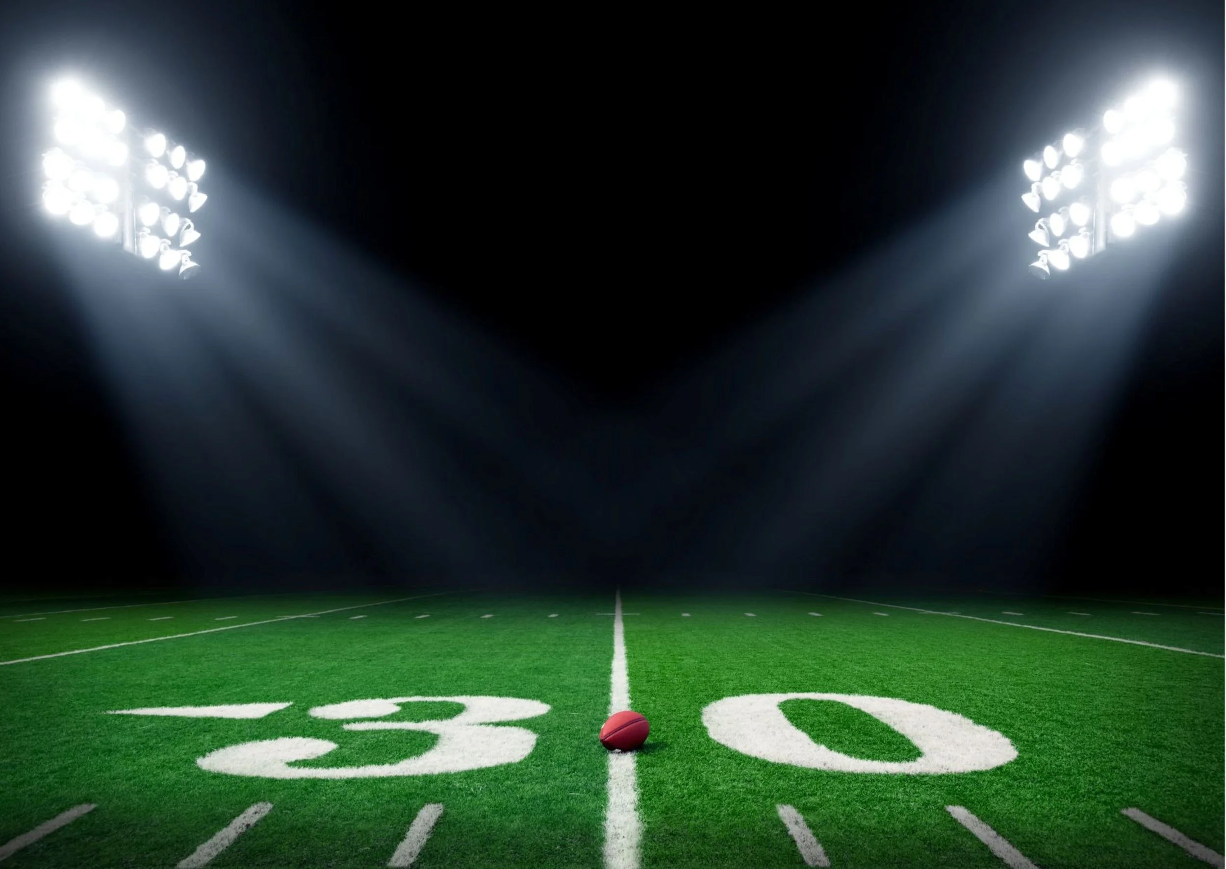 A football field lit by bright stadium lights, with a football centered on the 20-yard line.