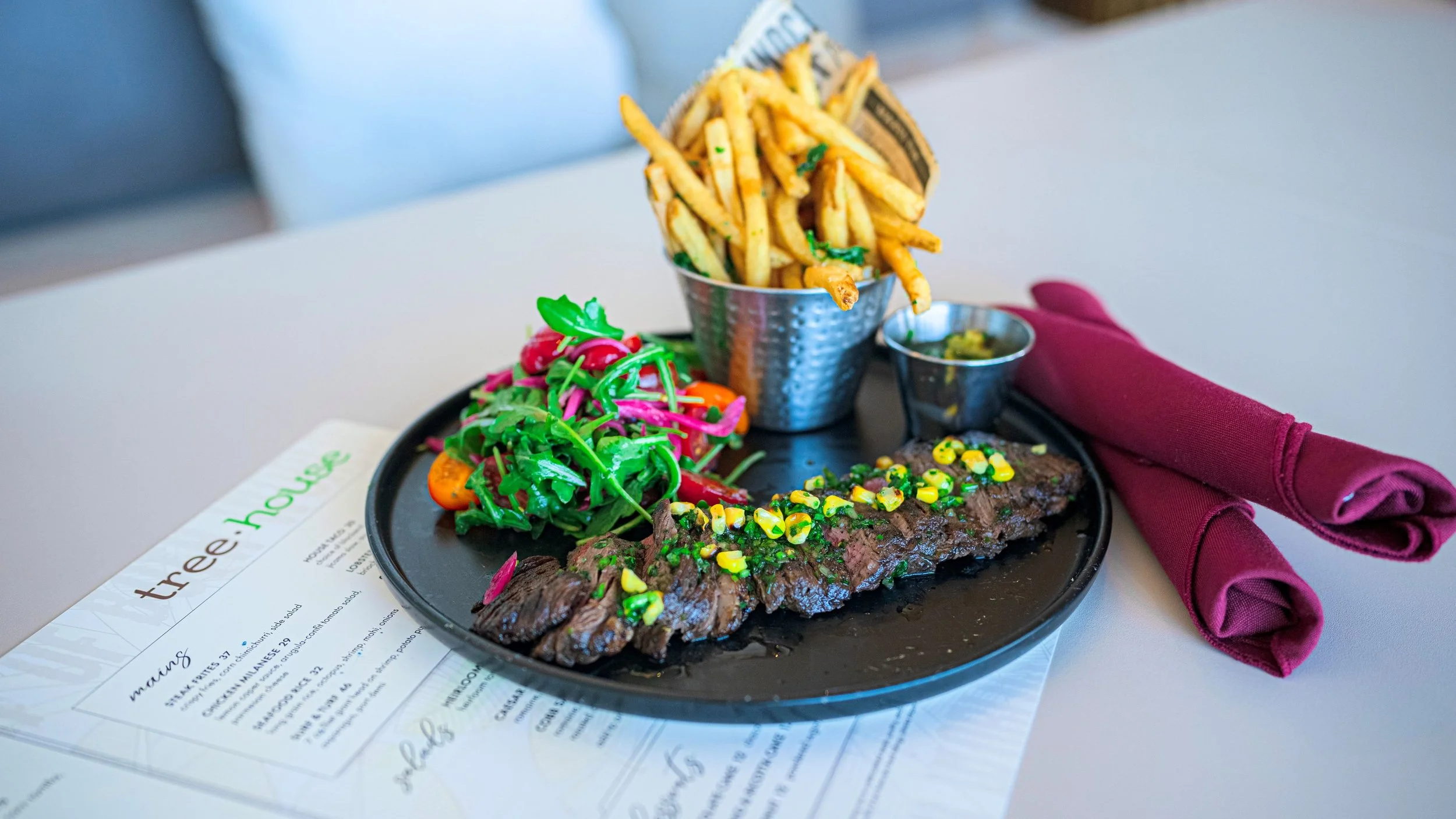 Steak with corn and scallions, side salad, French fries in a cup, small container of sauce, maroon napkin, and menu on white table.