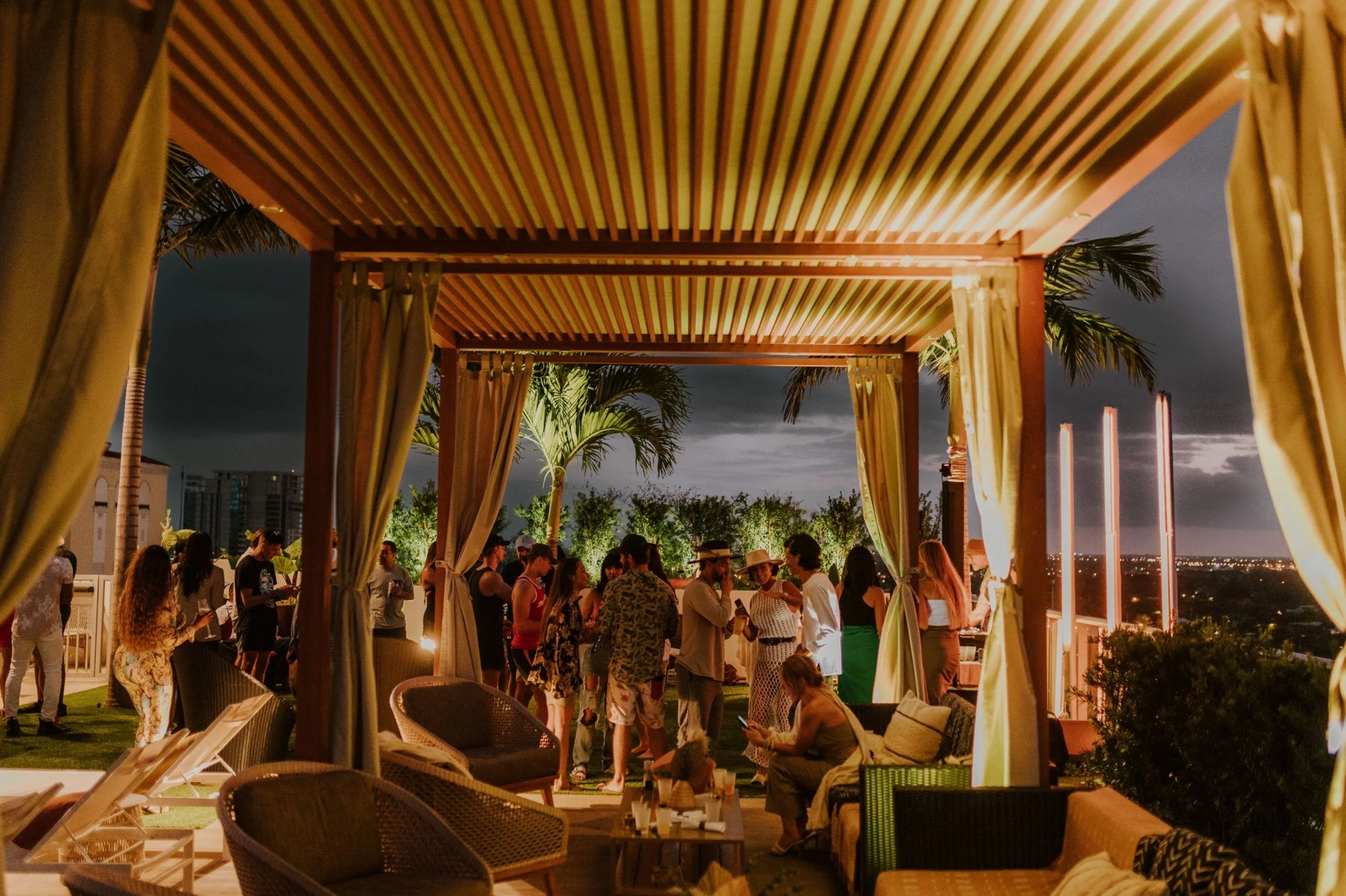 People enjoying a nighttime gathering on an outdoor patio with cabanas, palm trees, and city lights in the background.