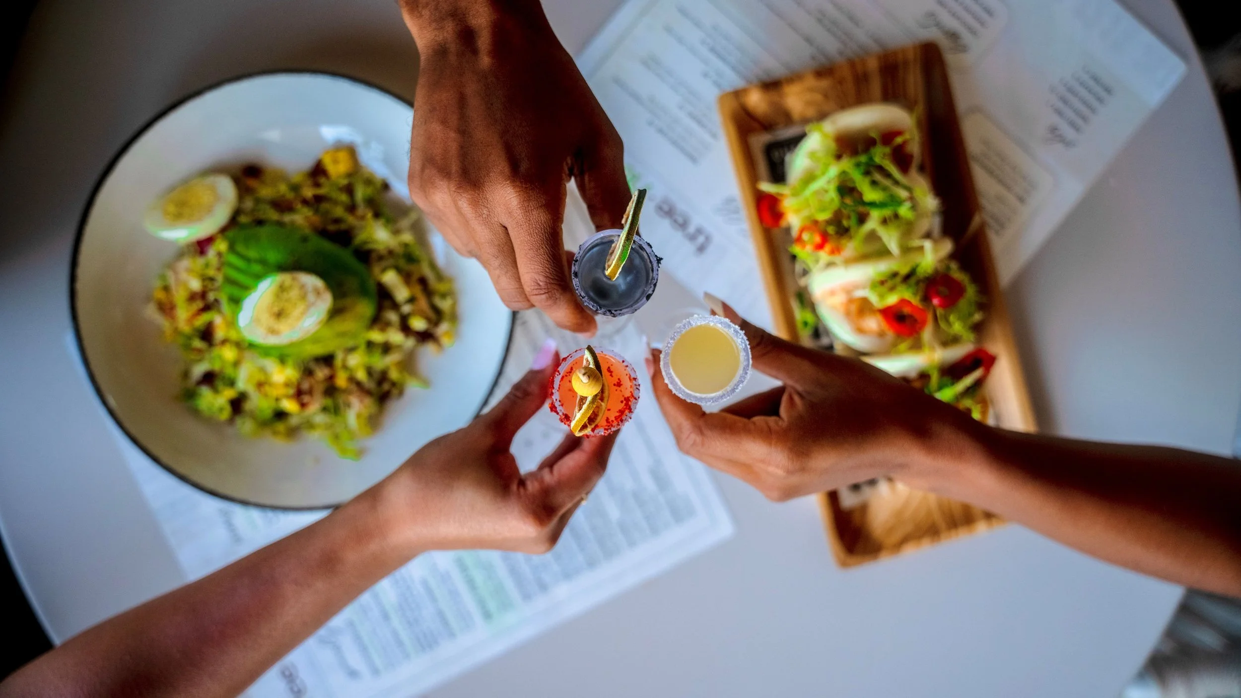 Three people clinking glasses of cocktails over a table with salad and tacos.