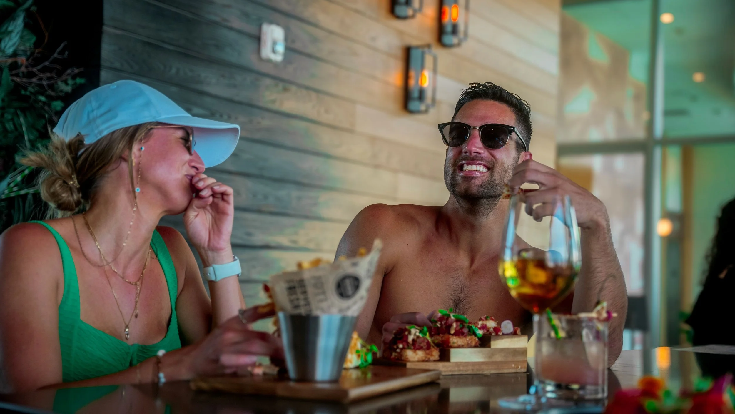 A woman and a man enjoy a meal together at a restaurant, laughing and smiling, with drinks and food on the table.