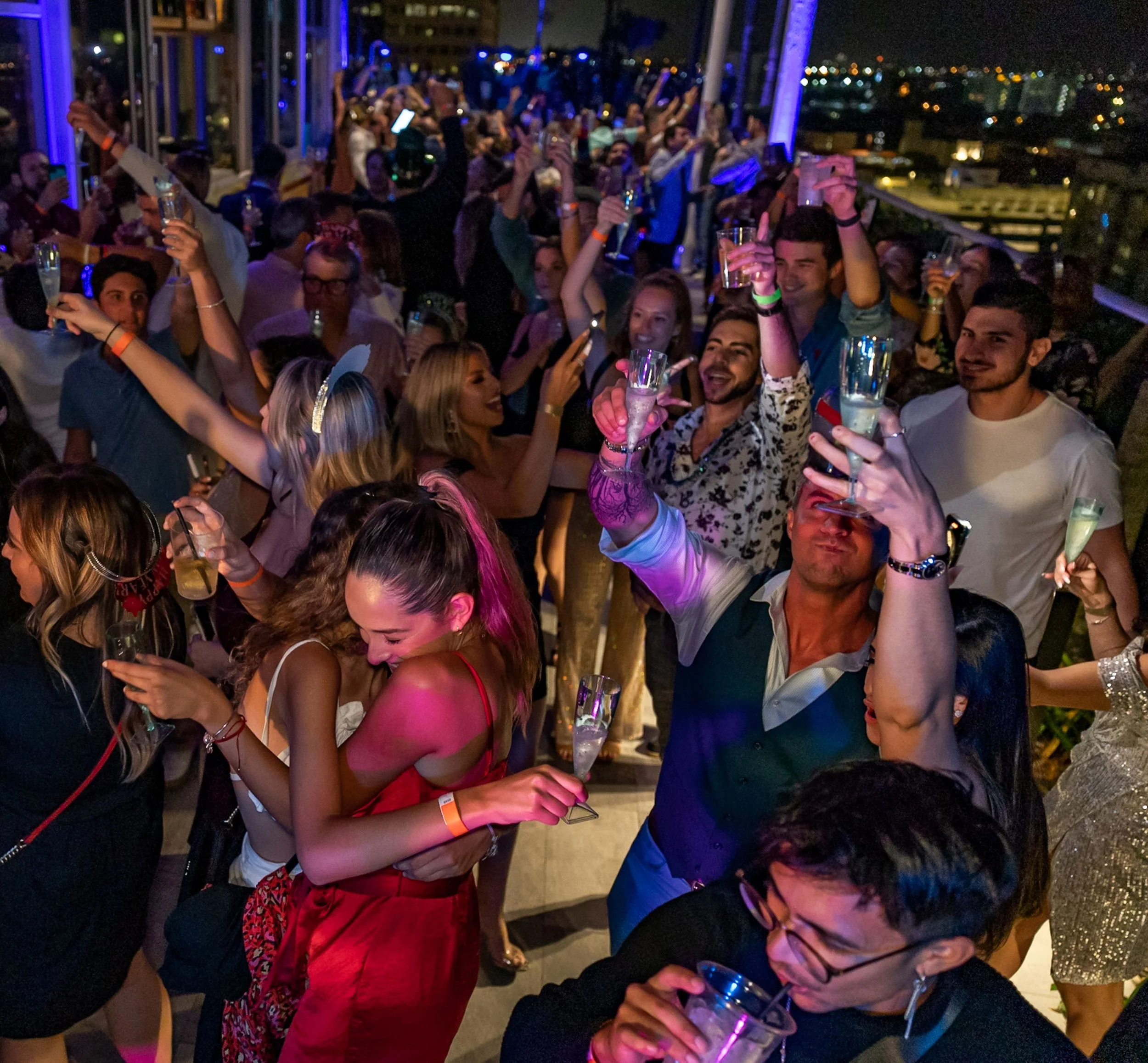 People dancing and celebrating at a lively nighttime party on a balcony with city lights in the background, holding drinks and raising their glasses.