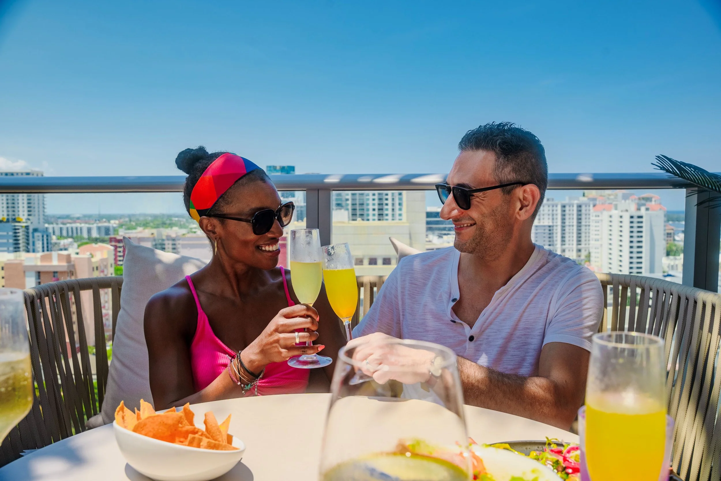 A man and woman on a rooftop terrace, holding champagne glasses, smiling, with a city skyline in the background. The woman has dark hair in a bun, wearing sunglasses and a pink top. The man has short dark hair, wearing sunglasses and a white shirt. The table has drinks, chips, and an appetizer.