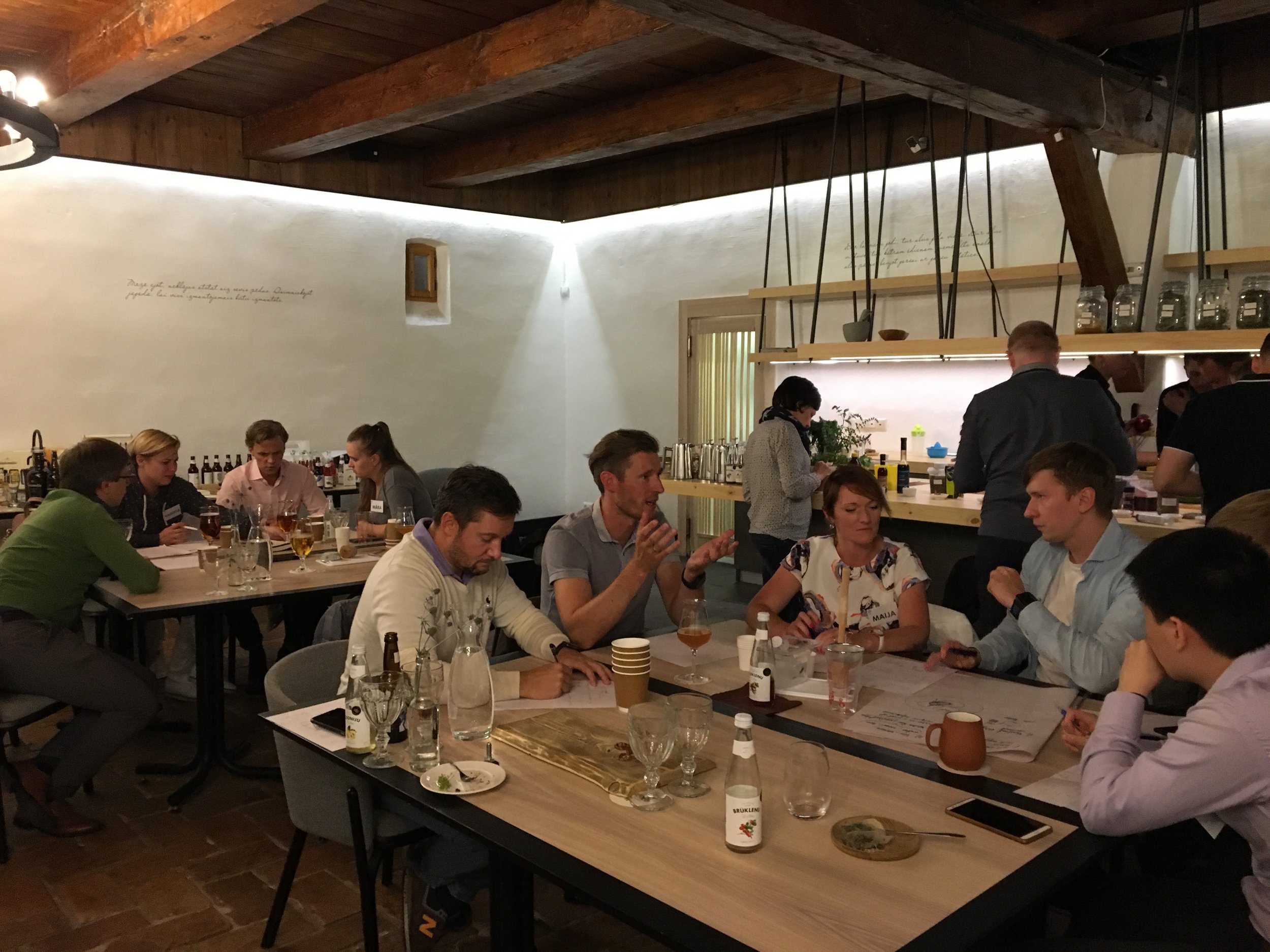 People gathering and talking at a restaurant with a bar in the background, bottles on shelves, and a white wall with written text.
