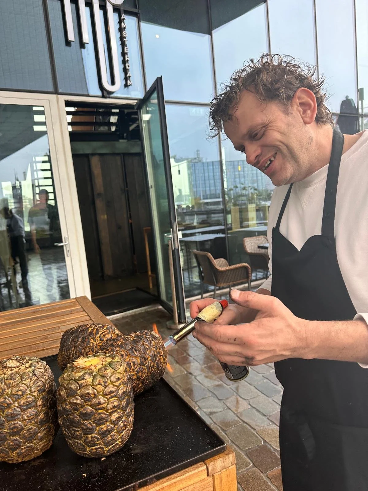 A man wearing a black apron is roasting pineapples with a culinary blowtorch at an outdoor restaurant patio.