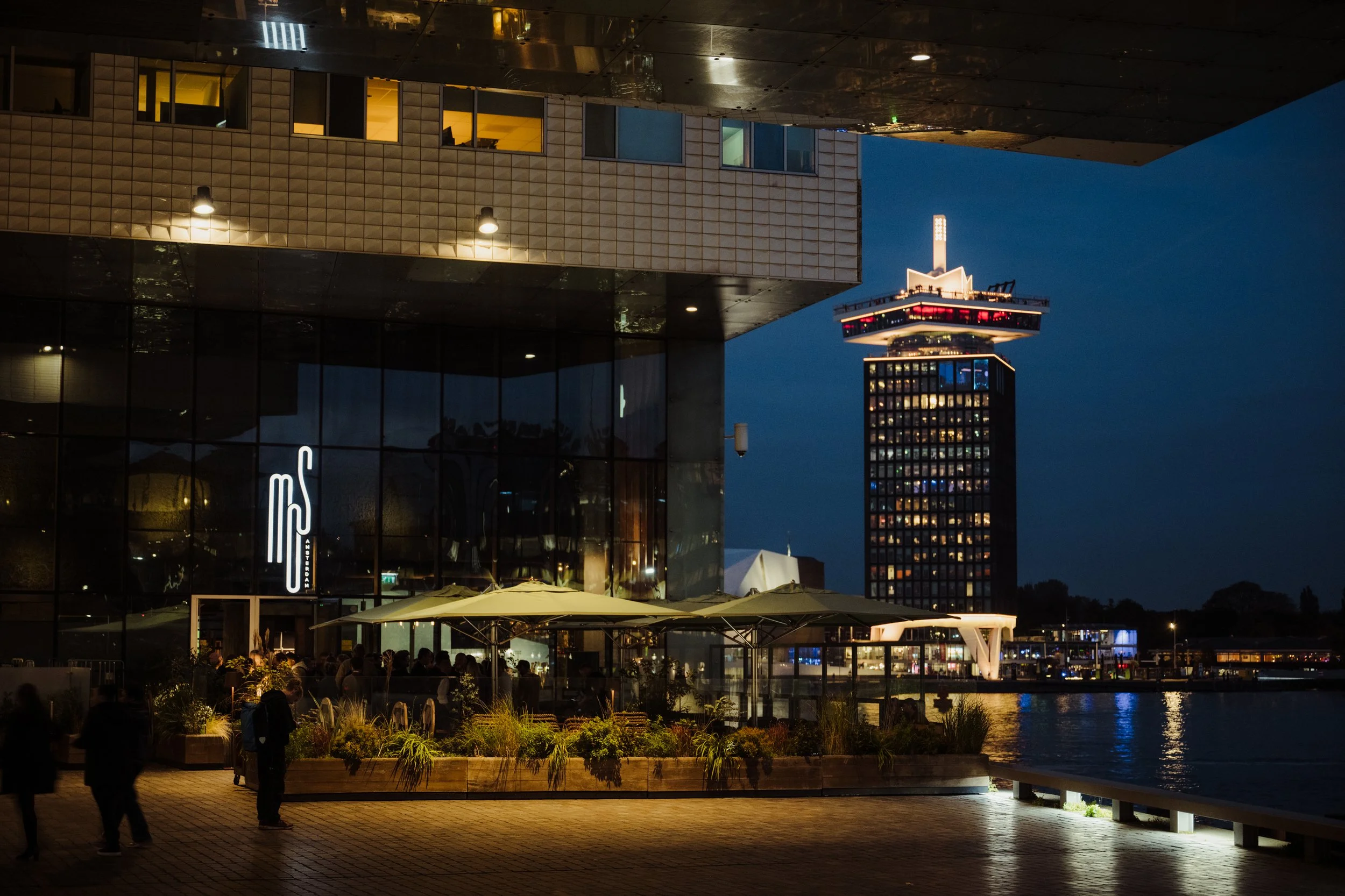 Night view of a modern waterfront building with people gathered on the patio, illuminated by exterior lights, and a tall lit tower in the background.