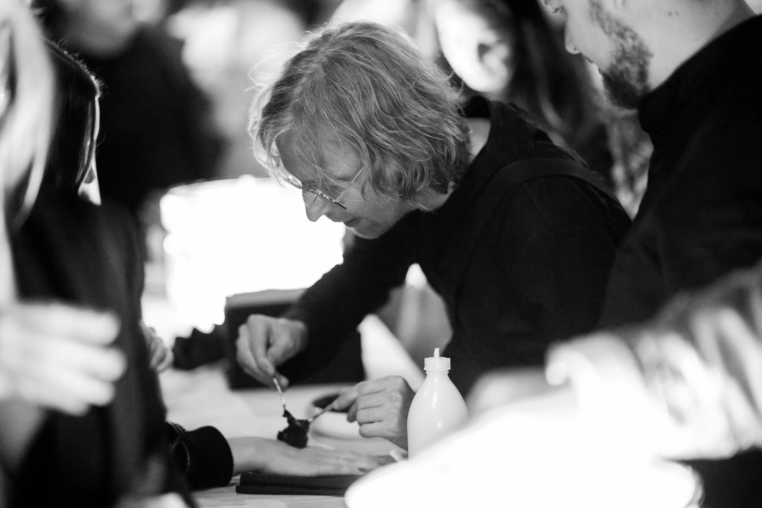 A person with glasses and wavy hair leaning over a table, applying black ink with a brush or tool. Several people are gathered around, observing or participating in an activity, in a well-lit environment.