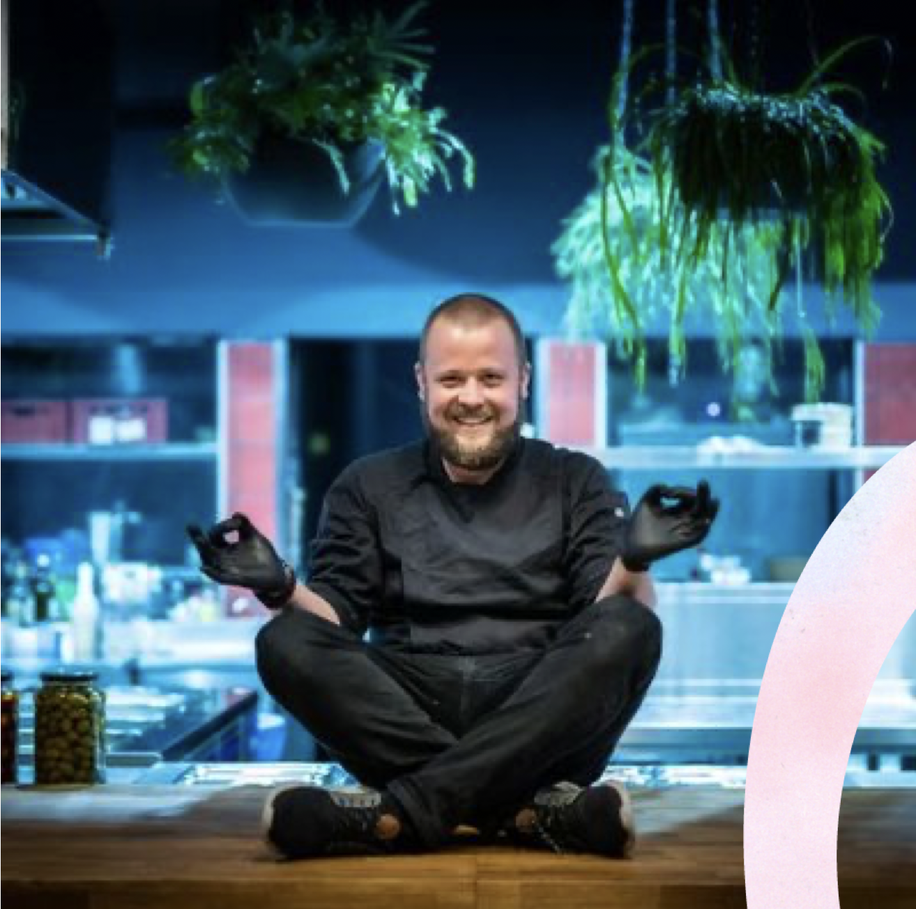 A smiling man sitting cross-legged on a wooden surface in a modern kitchen, wearing a black chef's coat and black gloves, with green plants hanging from the ceiling in the background.