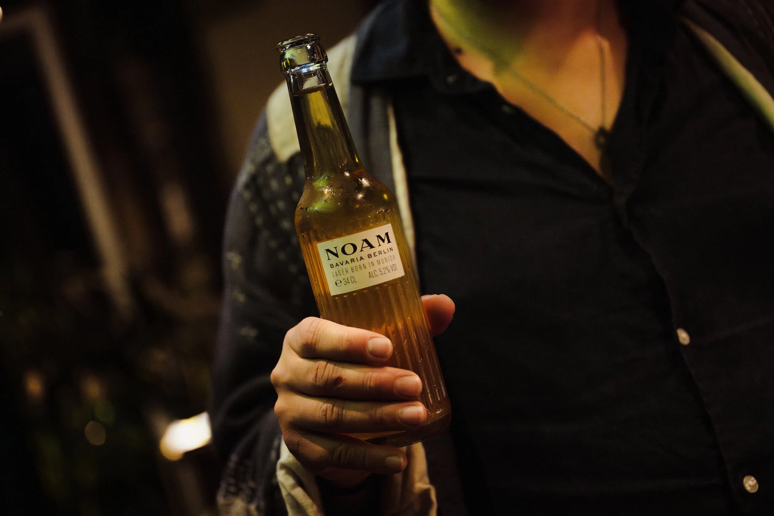 A person holding a bottle of NOAM Bavaria Berlin beer in a dimly lit setting.
