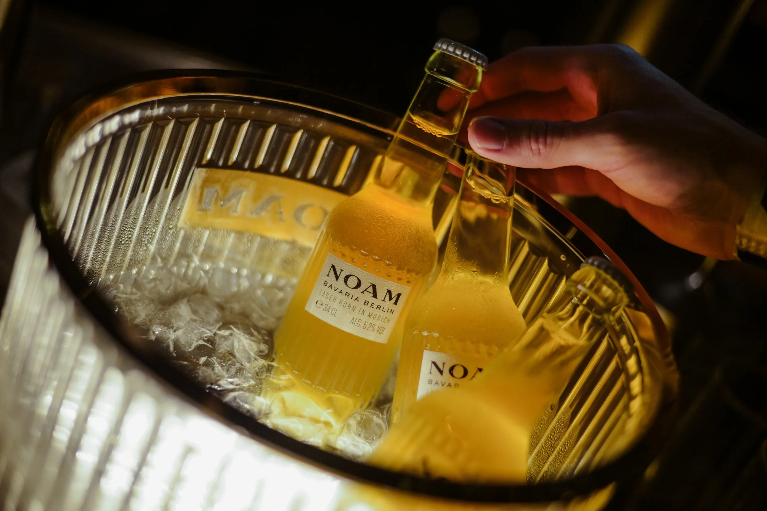 Two bottles of NOAM Berliner beer in ice bucket with ice cubes, hand reaching to pick up one bottle.