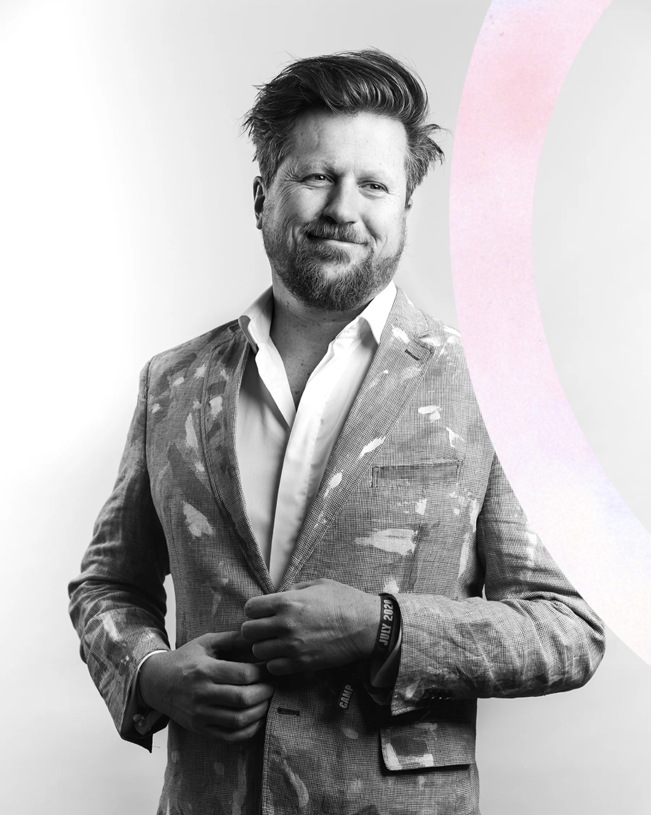 Black and white photo of a smiling man with a beard and styled hair, wearing a patterned blazer over a white shirt, adjusting his blazer with one hand, against a plain background.