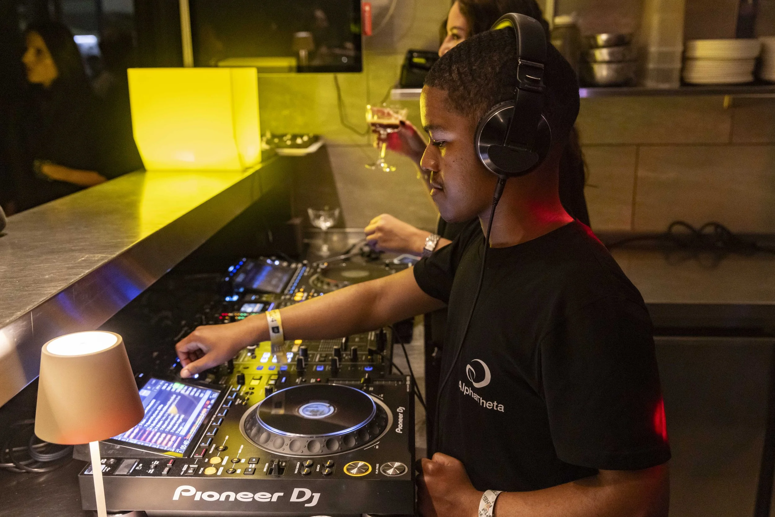 A young man DJing at a nightclub, wearing headphones and a black T-shirt with the word 'alphaheta' on it, with music equipment and a colorful light display in the background.