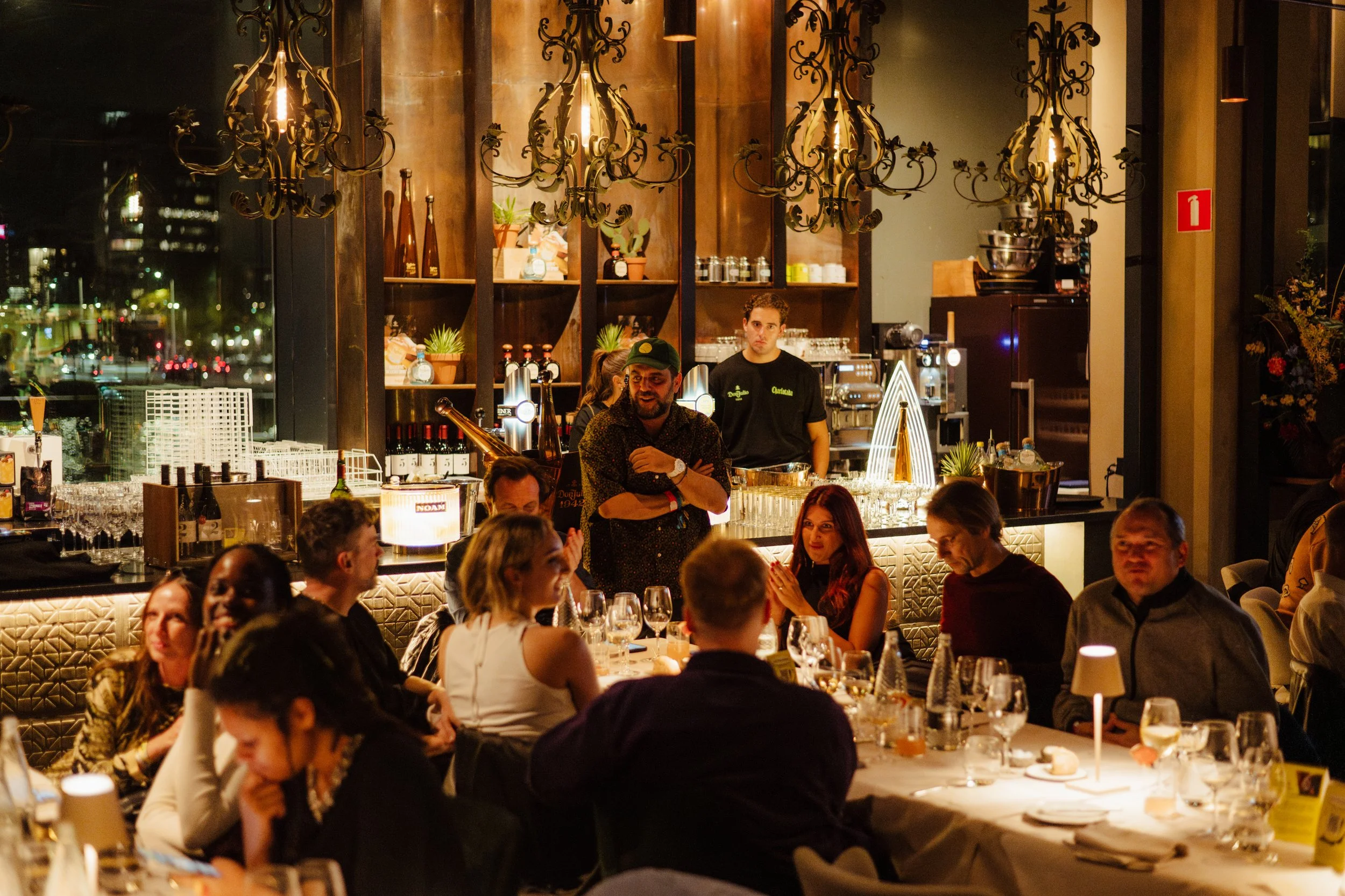 People dining at a restaurant with a bar in the background, dim lighting, and chandeliers.