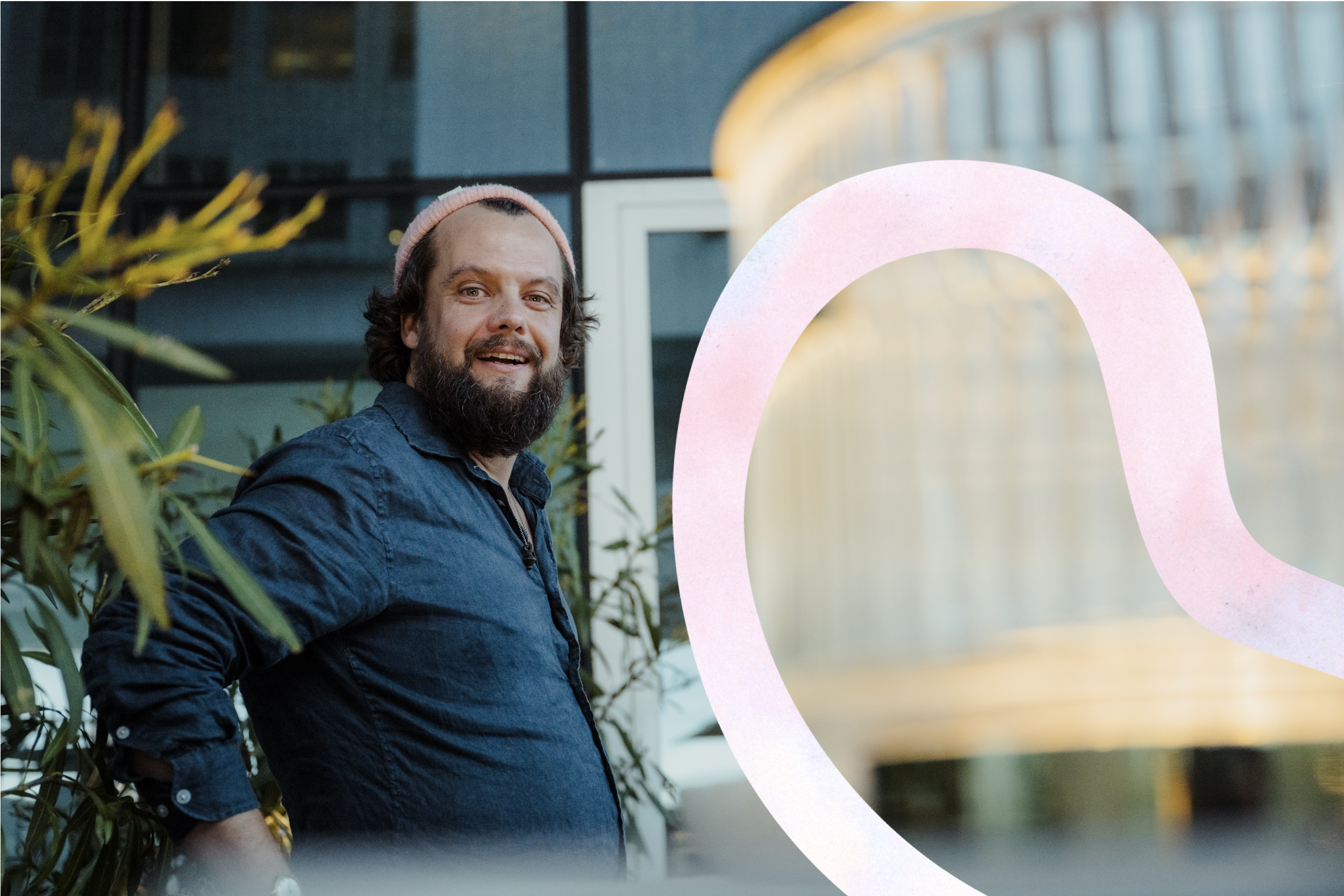 A man with a beard and curly hair, wearing a pink beanie and a dark blue shirt, standing outdoors near some green plants, smiling at the camera with a contemporary building with large glass windows in the background.