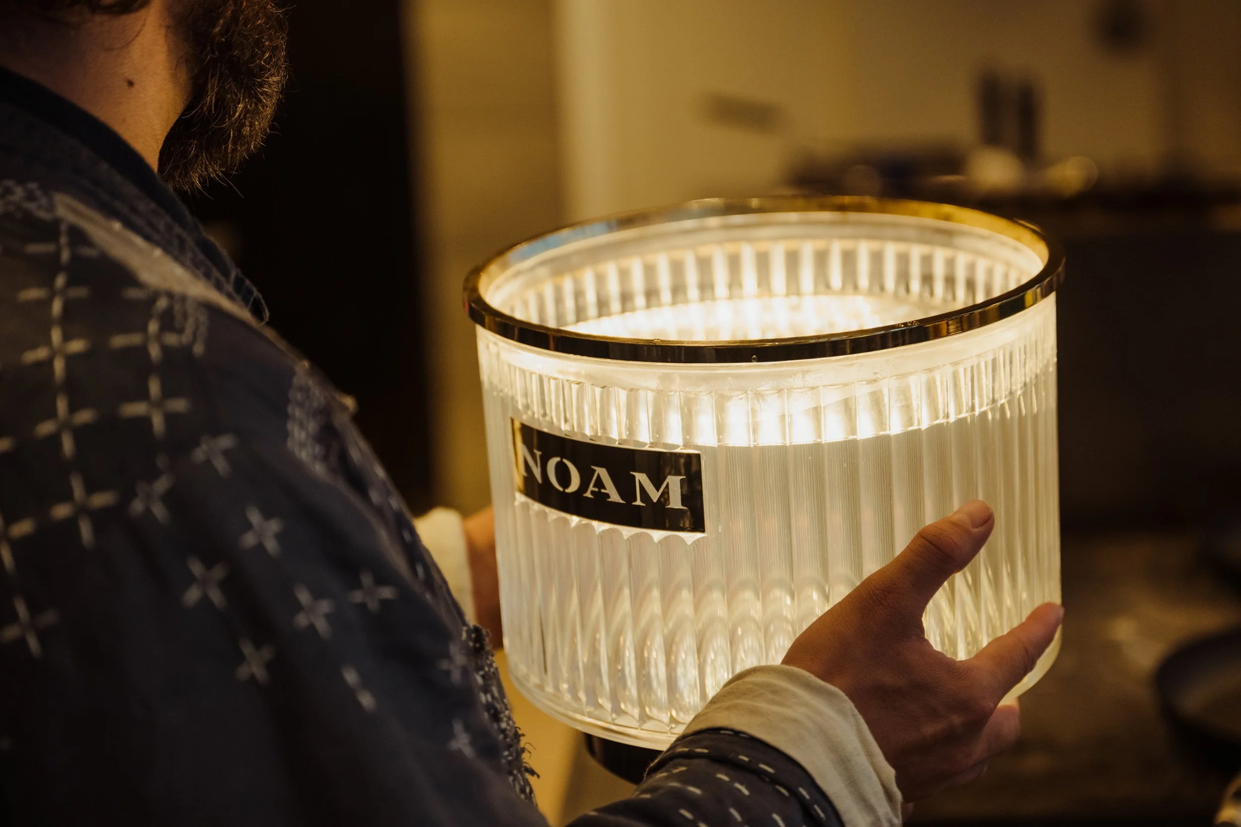 Person holding a lit, cylindrical lamp with the brand name 'NOAM' on it, in a dimly lit room.