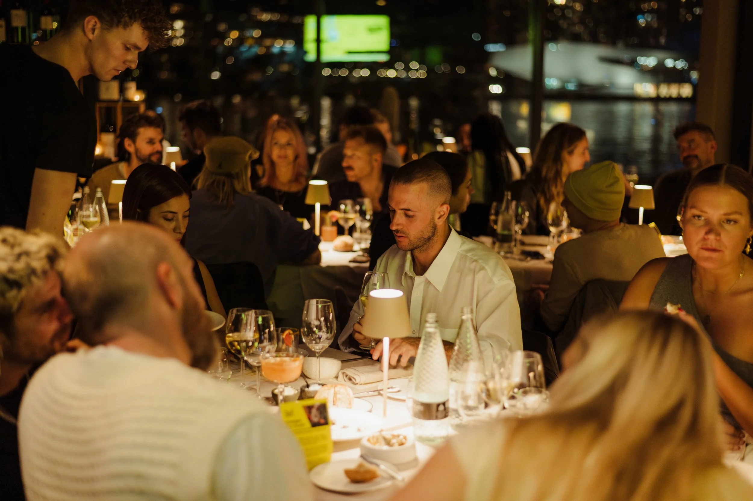 People dining at a restaurant during the night, with a view of city lights and water outside. The table is set with glasses, bottles, and plates, and a waiter is serving. The atmosphere is lively and warm.