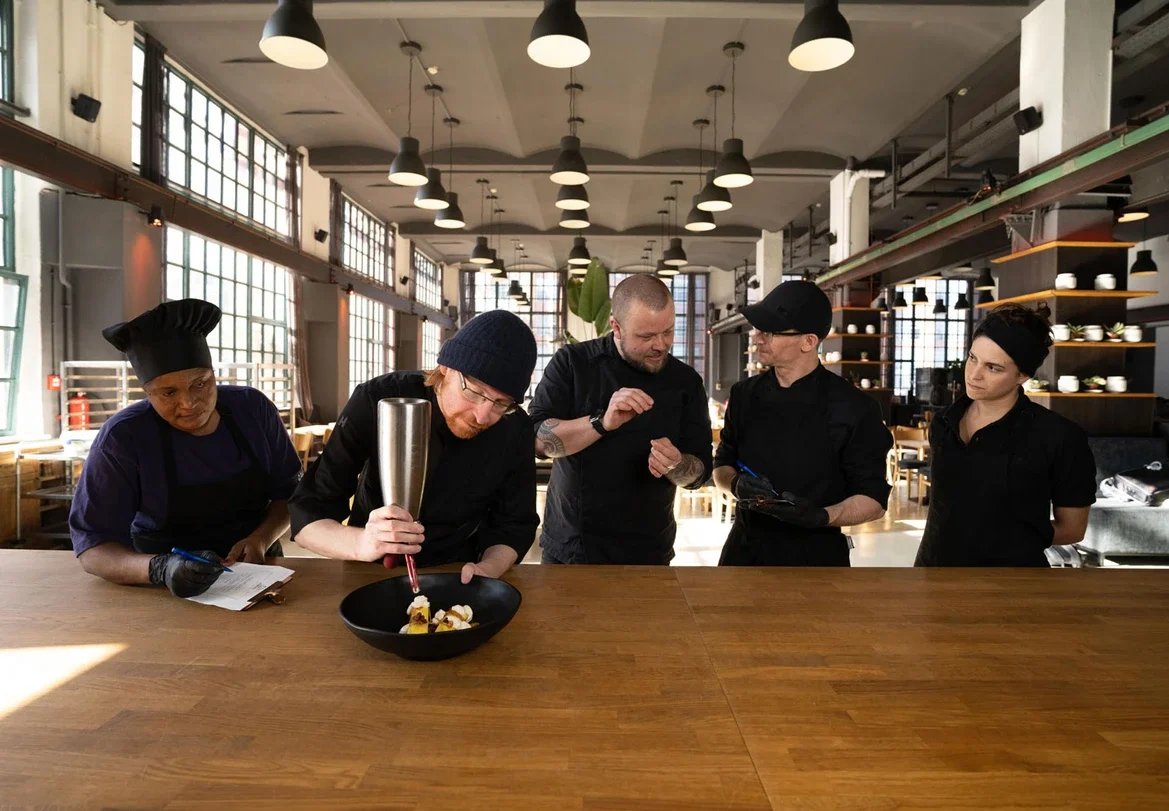 Five chefs in black uniforms and hats working together in a modern restaurant kitchen, with large windows and industrial-style lighting.