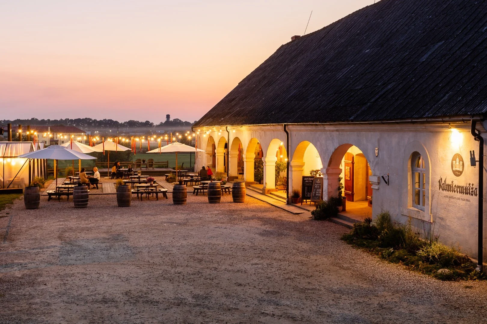 An outdoor patio of a restaurant or winery during sunset with tables, umbrellas, and barrel decorations, adjacent to a rustic building with arched doorways, string lights, and a few people dining.