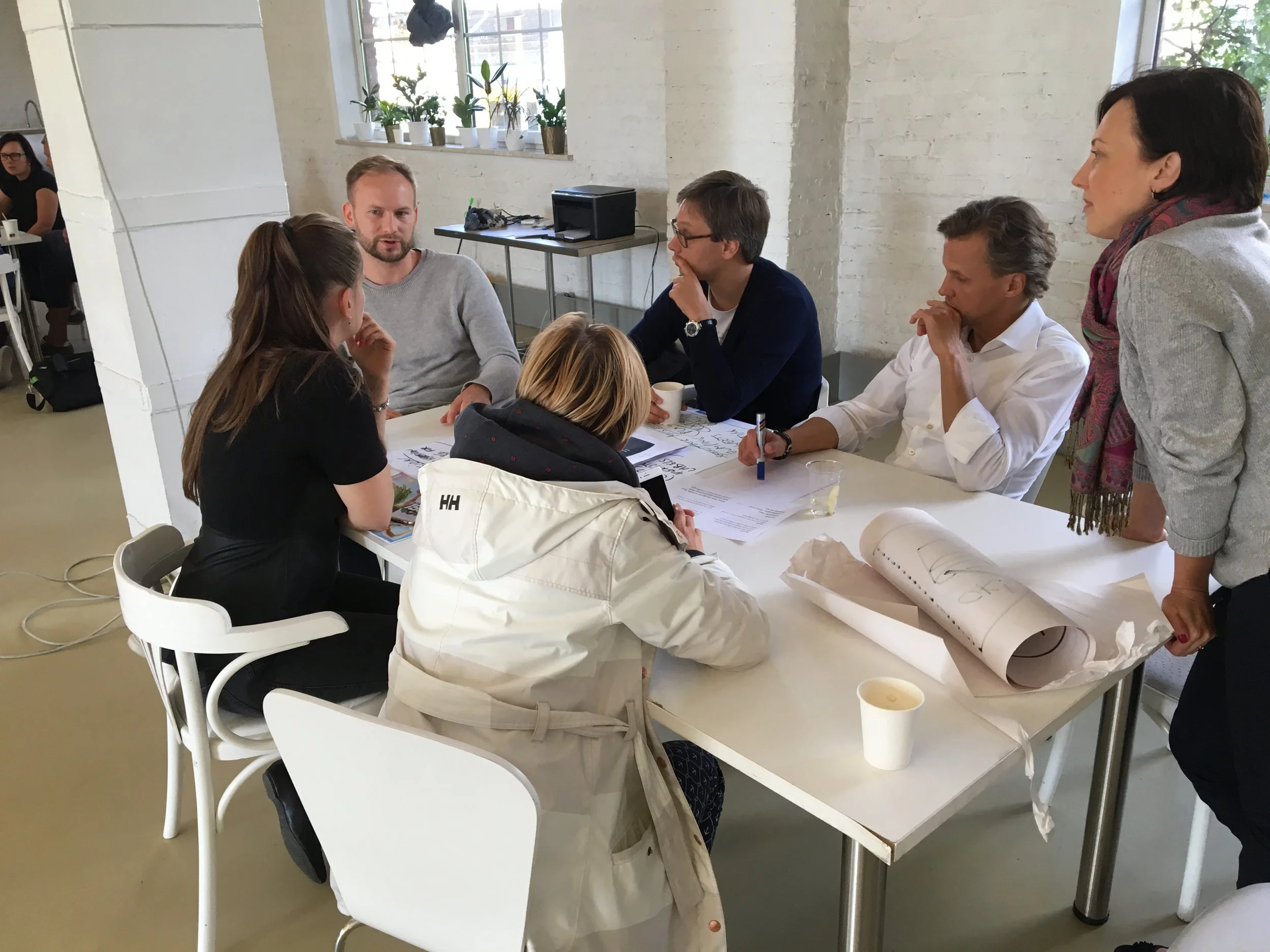 Group of six people sitting and standing around a table engaged in discussion in a modern office space with white walls, large windows, potted plants, and a person in the background.