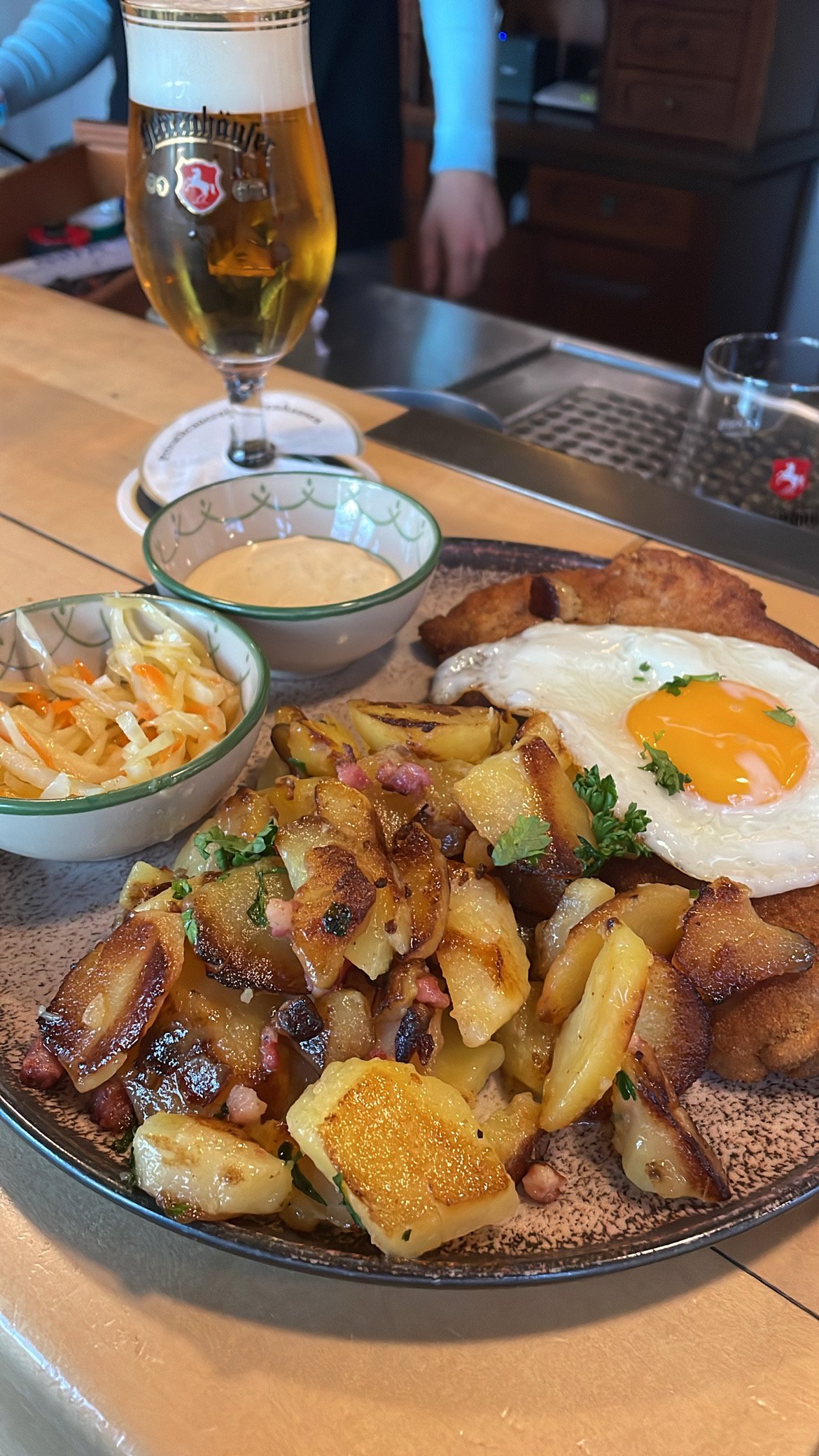 Ein Teller mit gekochten Kartoffeln, einem Spiegelei, Sauerkraut, und einem Stück Fleisch, dazu ein Glas Bier und zwei Saucenschalen mit Soße und Krautsalat auf einem Holztisch.