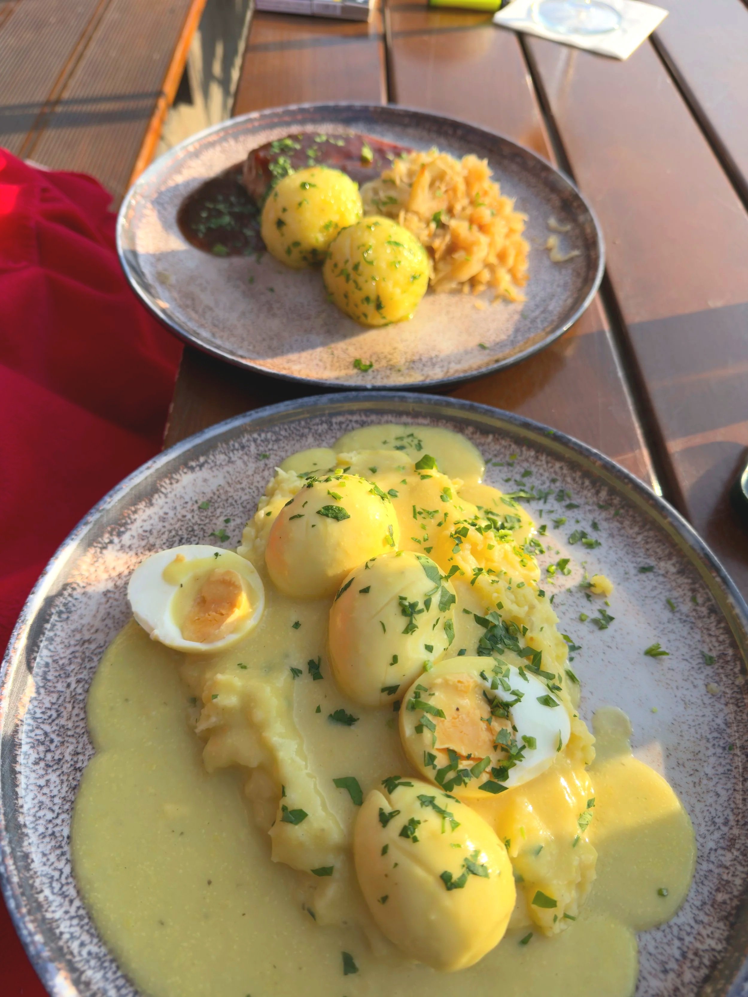 Ein Teller mit gekochten Eiern, einer hellgelben Soße und Kartoffelsalat, garniert mit frischer Petersilie, auf einem Holztisch im Freien.