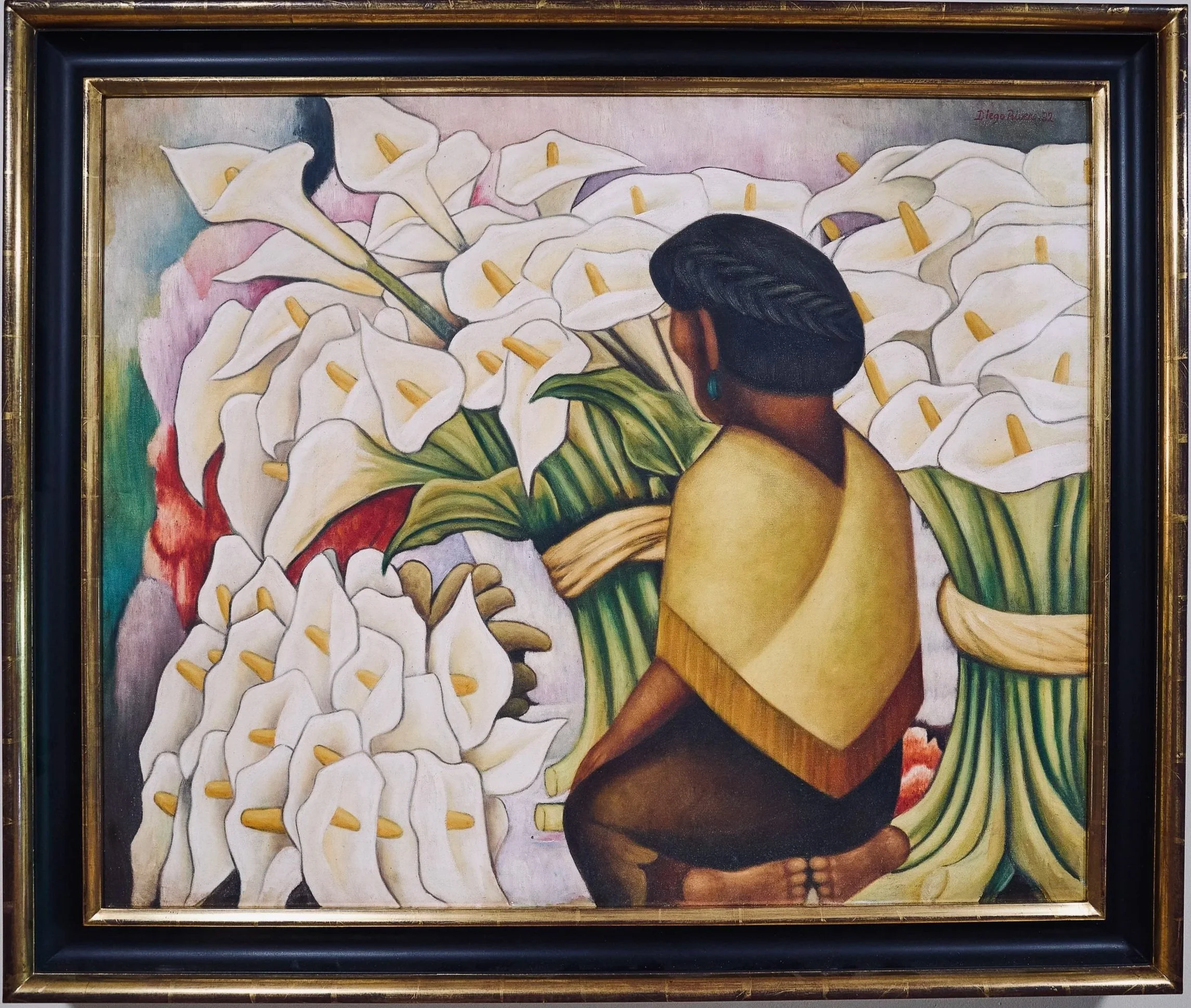 A painting of a woman with dark hair and dark skin, wearing a green and yellow skirt and turquoise earrings, holding a large bouquet of white calla lilies with yellow centers. The painting is framed with a black and gold frame.