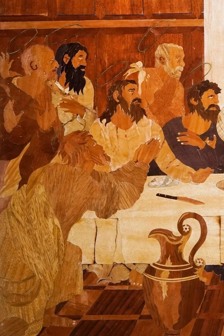 Wooden art depiction of Jesus Christ and his apostles during the Last Supper, with some figures standing and others sitting at a table, in a warm brown tone.