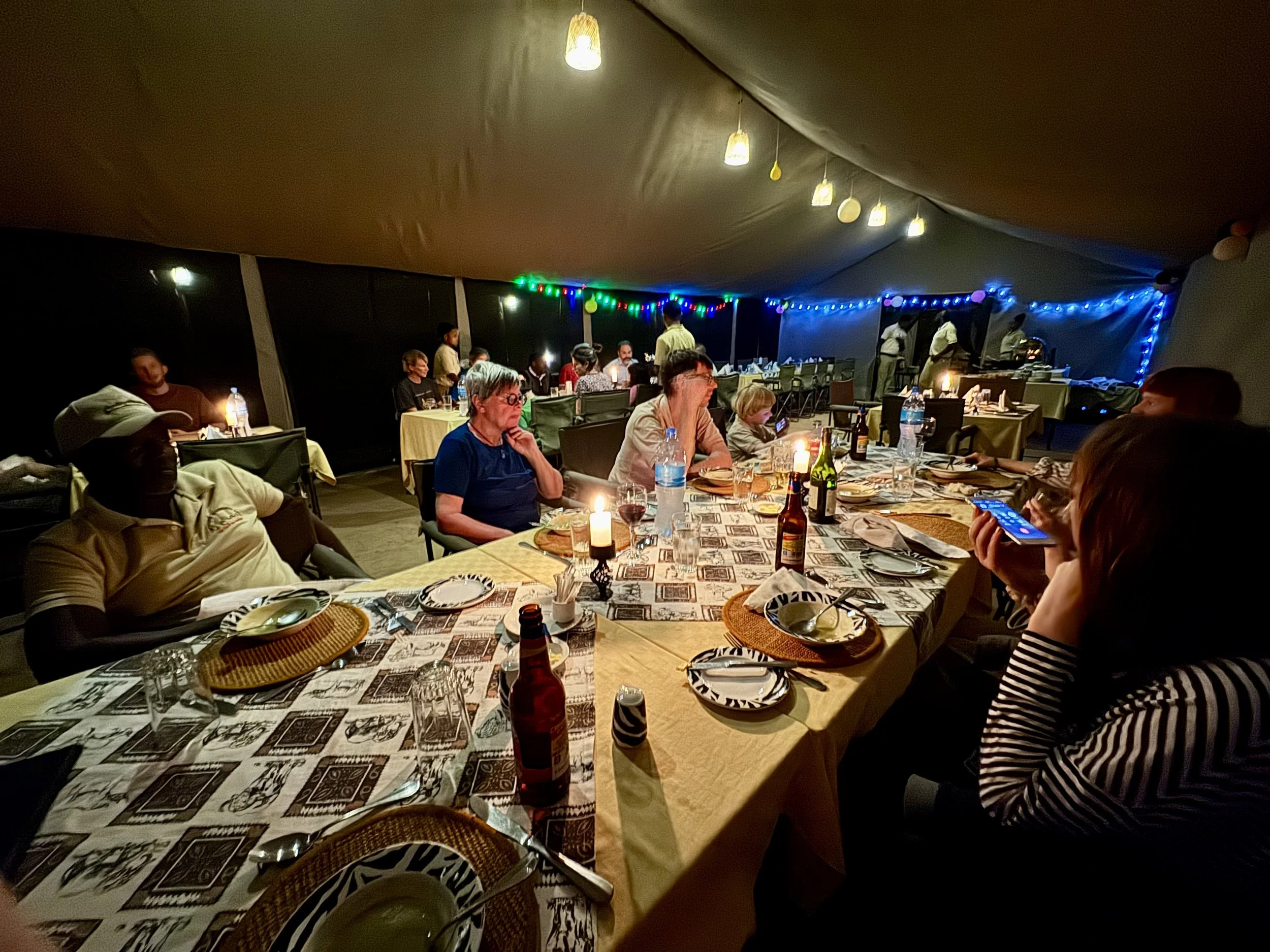 People sitting around a dining table inside a tent at night, with decorated lights and a buffet area in the background.