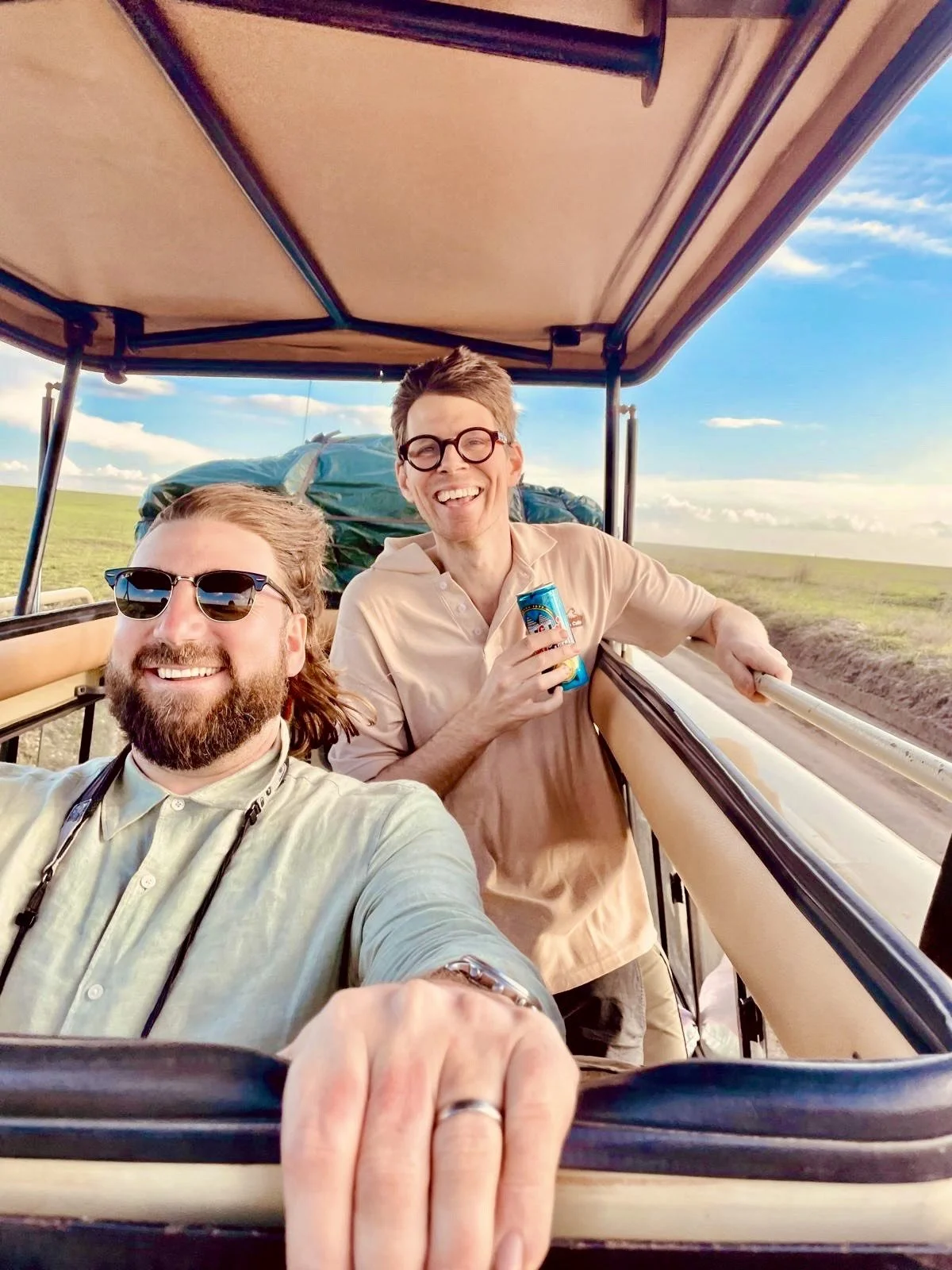 Two men riding in an open-air tram through a grassy landscape, smiling and taking a selfie. One is wearing sunglasses and a light shirt, the other is holding a beverage and wearing glasses and a beige shirt. The sky is blue with some clouds.