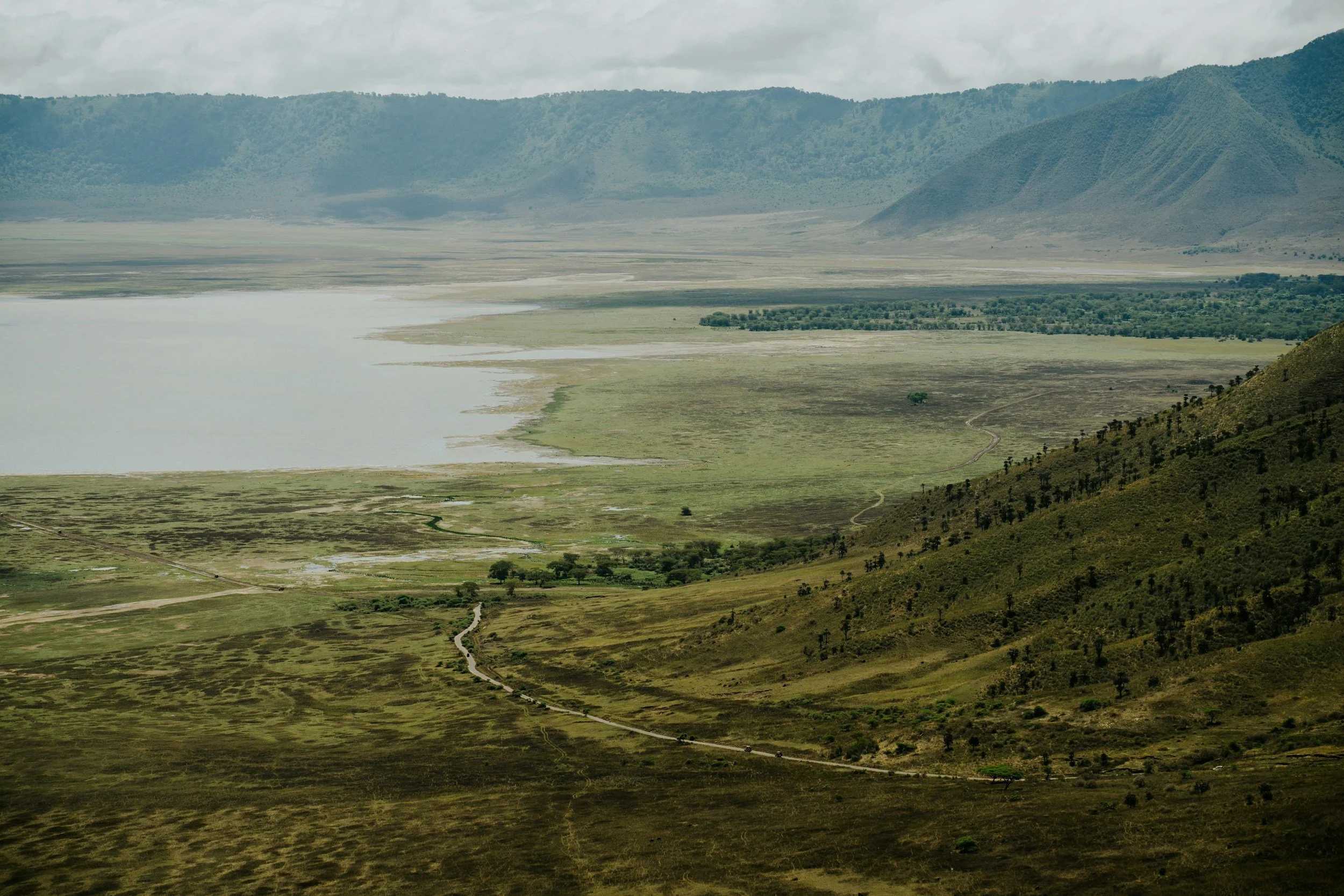 A vast landscape with a lake, green grassy plains, hills, and mountains in the background under a cloudy sky.