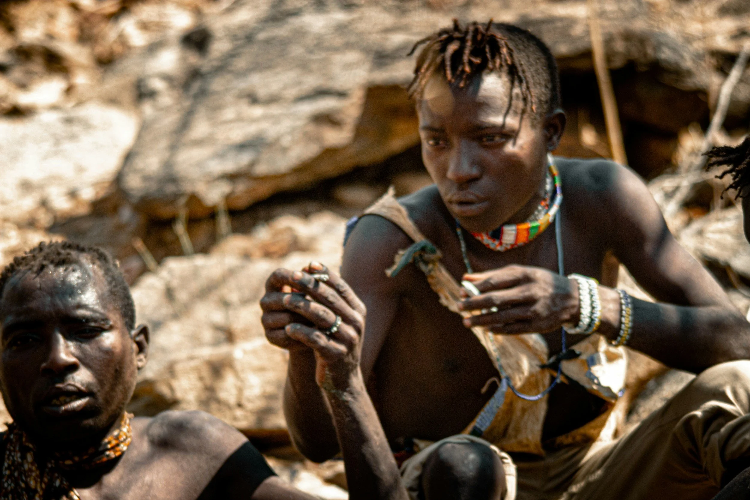 Two individuals from an African tribe with traditional attire and jewelry, sitting outdoors near rocks.