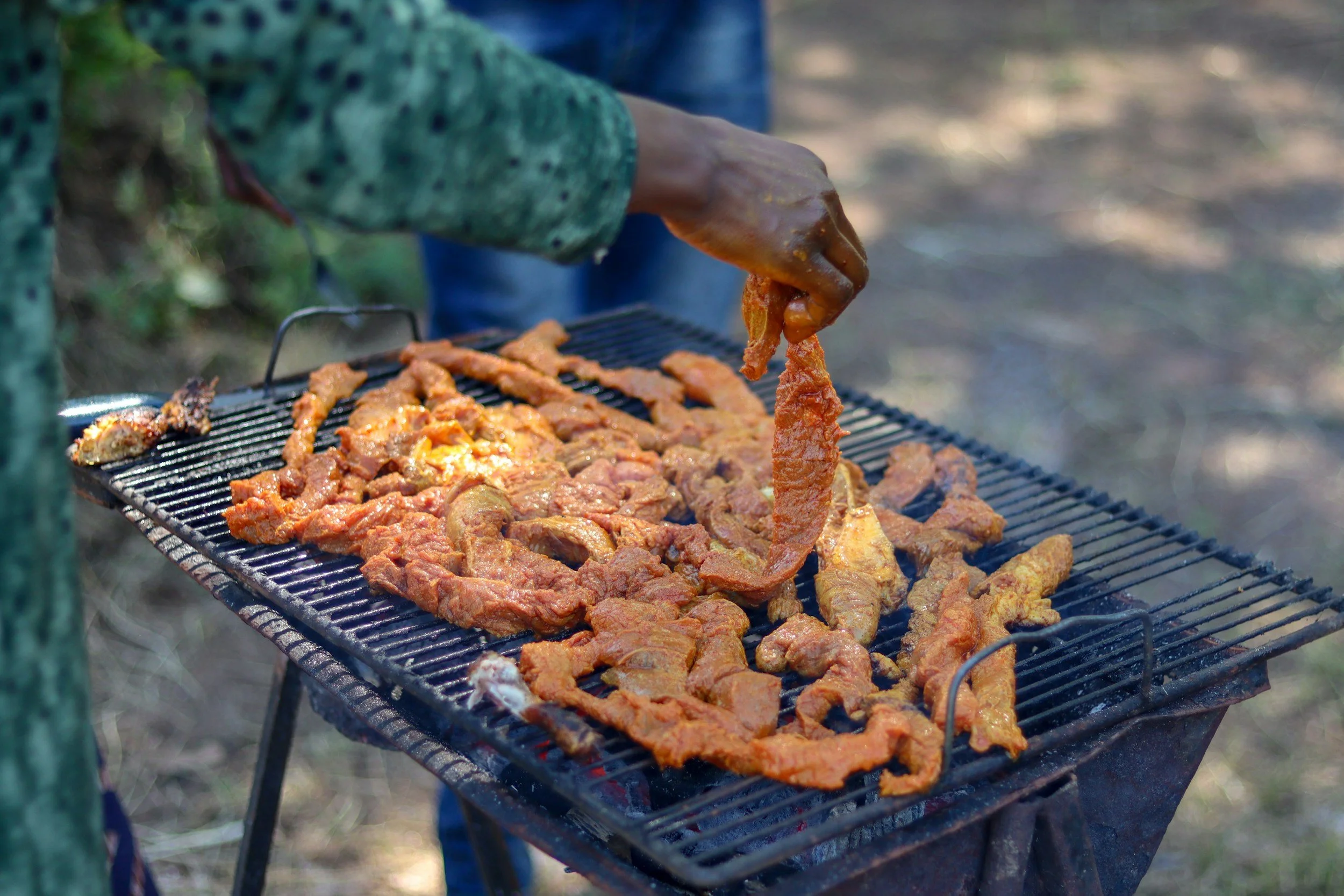 Person grilling marinated meat on a barbecue grill outdoors during the daytime.
