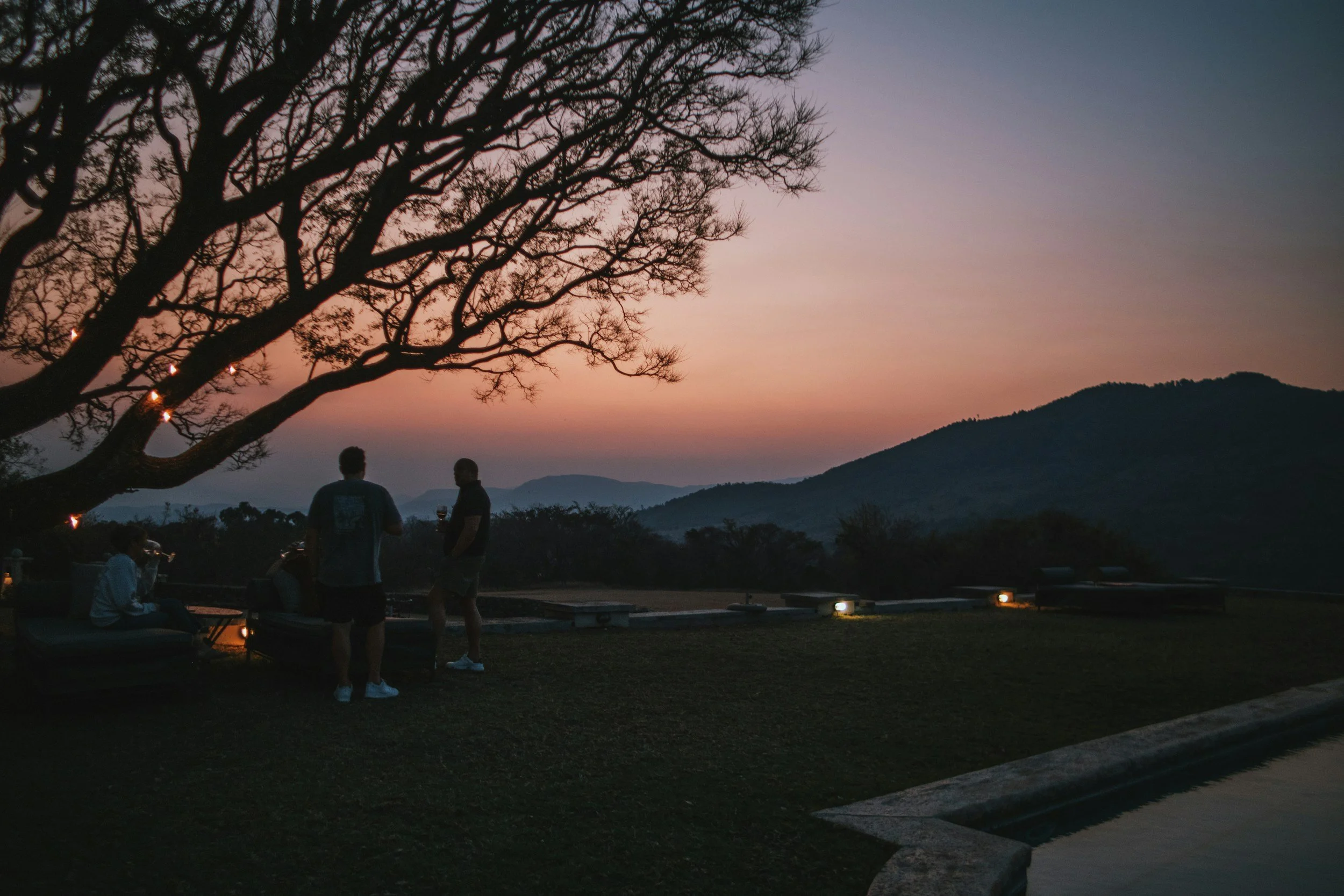 People gathering outdoors at sunset with mountains in the background, a large tree and some benches, with ambient lighting on the ground.