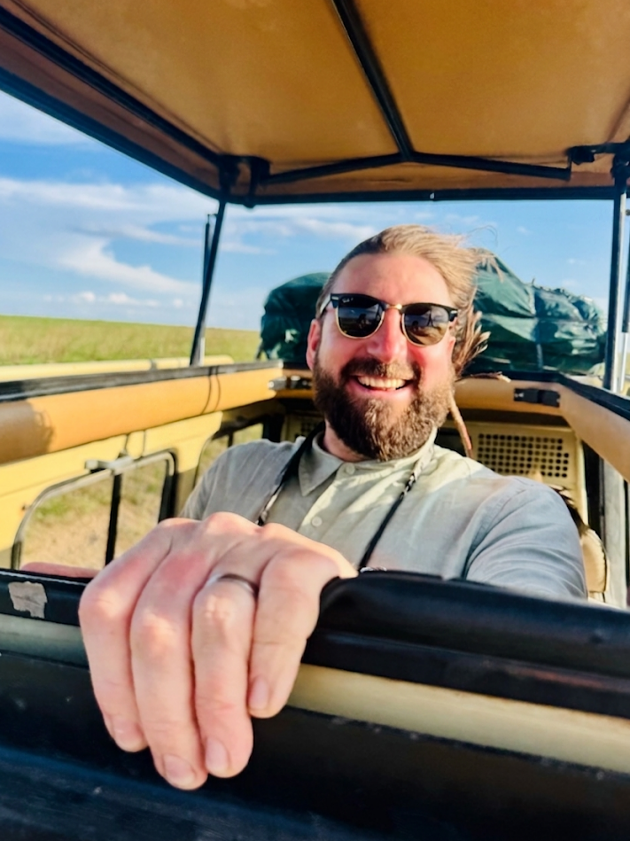 Smiling man with sunglasses taking a selfie from a hot air balloon basket during a sunny day, with open sky and grassy landscape in the background.