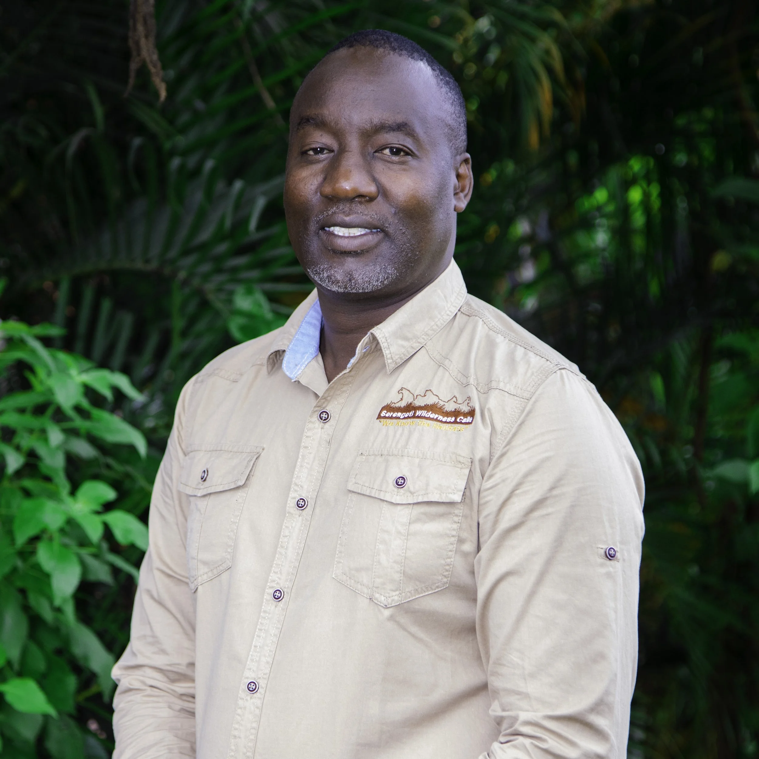 A man standing outdoors in front of lush green foliage, wearing a beige shirt with a logo on the chest area.
