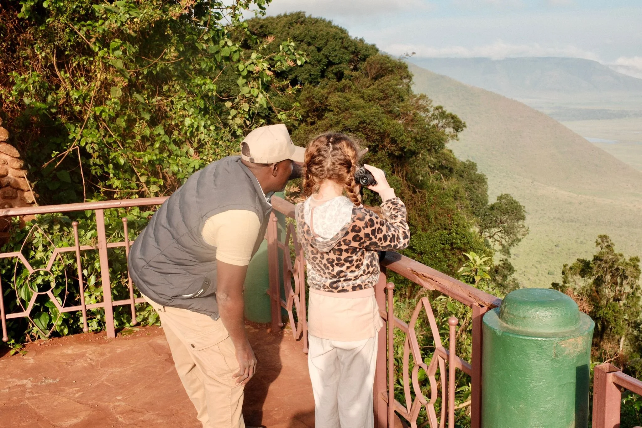Two people, a man and a girl, are looking through binoculars at a mountain landscape from a viewing platform surrounded by greenery.