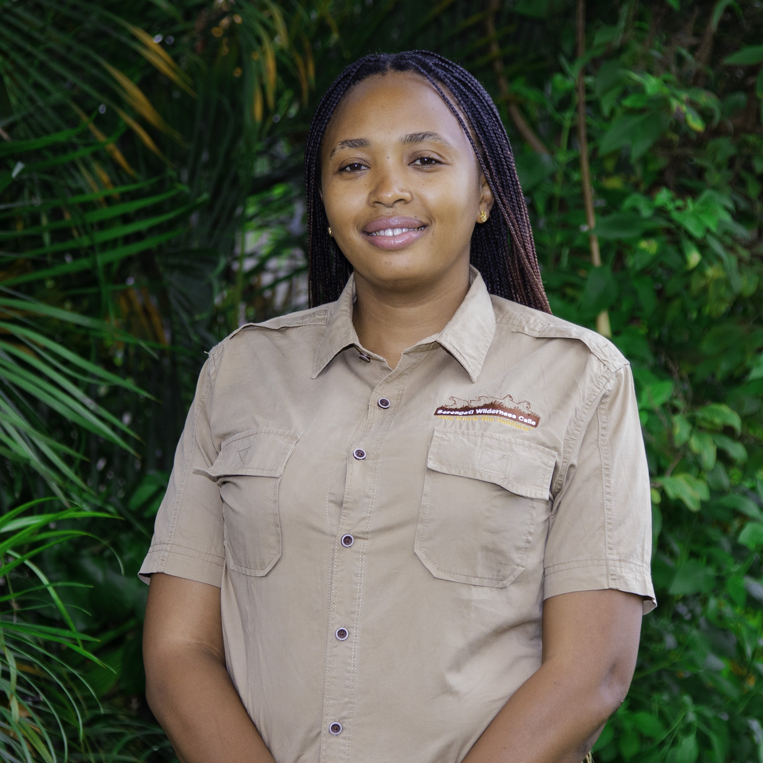 A woman wearing a beige safari shirt with a wilderness logo, standing outdoors in front of green tropical plants, smiling gently.