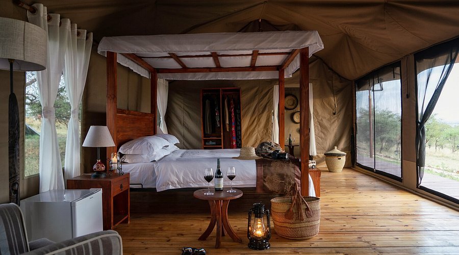 Interior of a safari tent bedroom with a wooden four-poster bed, white bedding, bedside tables with lamps, glasses of wine on a small round table, wicker baskets, and large windows with curtains showing an outdoor landscape.